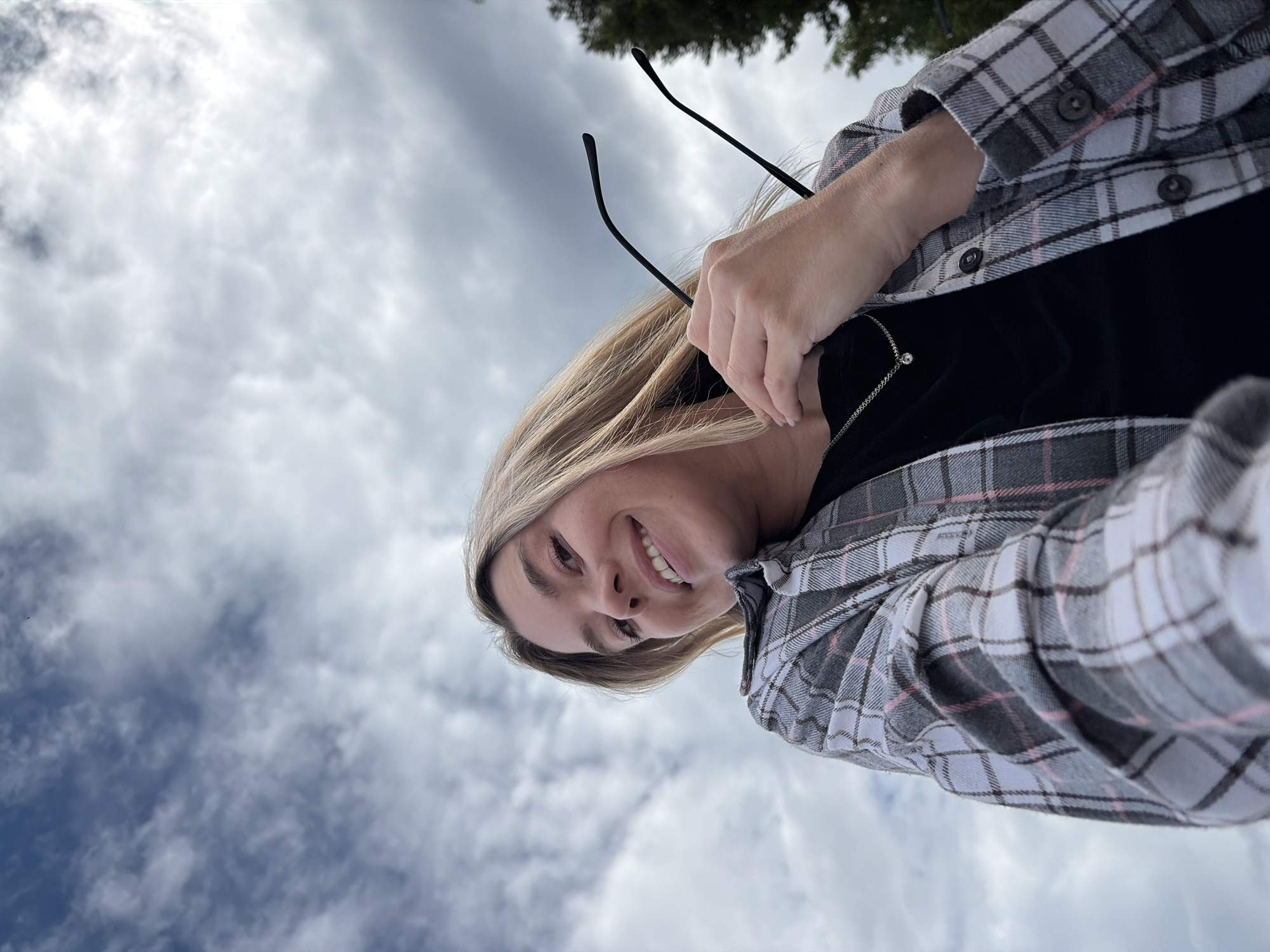 A low-angle, outdoor selfie shows a young woman with blonde hair and a bright smile looking up at the camera. She is wearing a black top under a grey and white plaid shirt with pink and dark grey accents. She holds a pair of black-rimmed glasses in her right hand, bringing them up towards her face. A delicate silver necklace with a single pendant is visible around her neck.
The background is dominated by a cloudy sky, with dark grey and white clouds suggesting overcast weather. The bottom of the image shows a hint of green foliage, possibly trees, against the sky, suggesting the photo was taken outdoors. The lighting indicates it is daytime, though the clouds suggest it may not be a particularly sunny day. The woman's cheerful expression and the casual attire imply a relaxed, possibly spontaneous moment. The provided location context of Chaikovsky, Russia, places the scene in an urban or semi-urban outdoor environment within that region. No visible text is present in the image.
