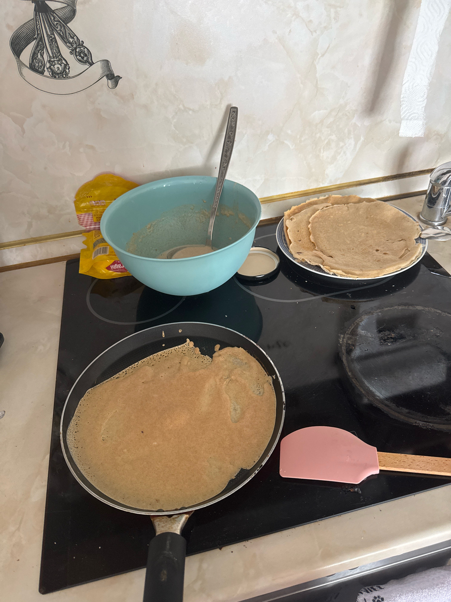 The image depicts a kitchen scene where crepes are being prepared. In the foreground, a black frying pan on a black stovetop contains a partially cooked crepe. To the right of the frying pan, a pink spatula with a wooden handle rests on the stovetop. Behind the frying pan, a plate with several cooked crepes is visible. To the left of the plate, a light blue bowl contains some batter and a silver ladle. A yellow package is also visible near the bowl. The background shows a marble-patterned backsplash and a roll of paper towels. The stovetop appears to be an electric induction cooktop. The overall impression is one of home cooking in progress. There are no visible indicators to suggest the location is Sofia, Bulgaria.