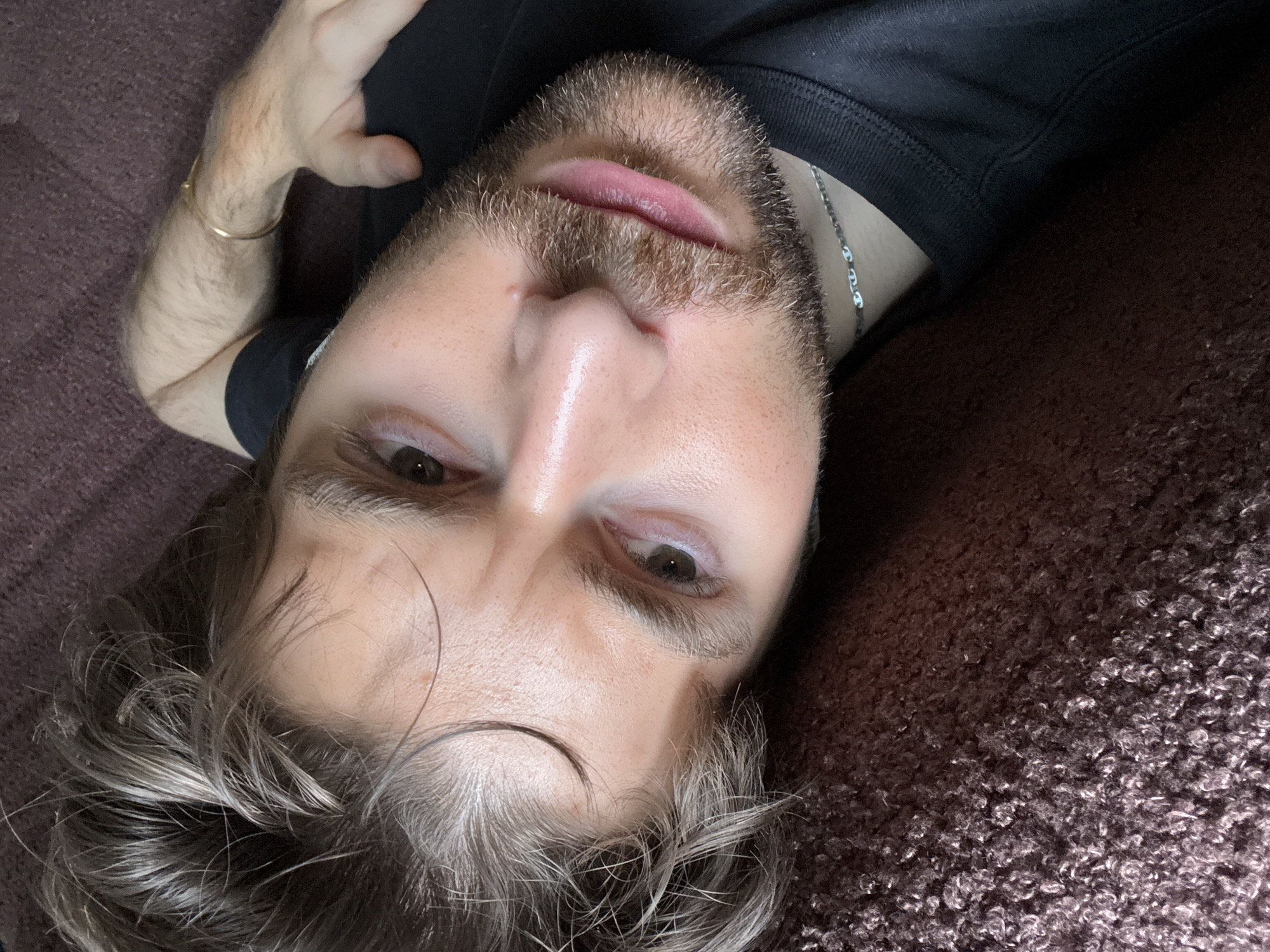 A close-up, high-angle portrait captures a person lying on their back on a dark reddish-brown, textured surface, possibly a sofa or carpet. The person looks directly into the camera. They have a short beard or stubble and wavy, dark hair with visible lighter or greyish strands, some of which fall across their forehead.
The person is wearing a black t-shirt or top. A thin silver chain necklace is visible around their neck, and a simple gold-colored bangle or bracelet adorns their left wrist. Their eyes are open, and their lips are slightly parted. The lighting appears soft, indicating an indoor setting with no strong direct light. There are no specific cues within the image to suggest a time of day or external weather. The person's expression is neutral and direct. No text is visible in the image.