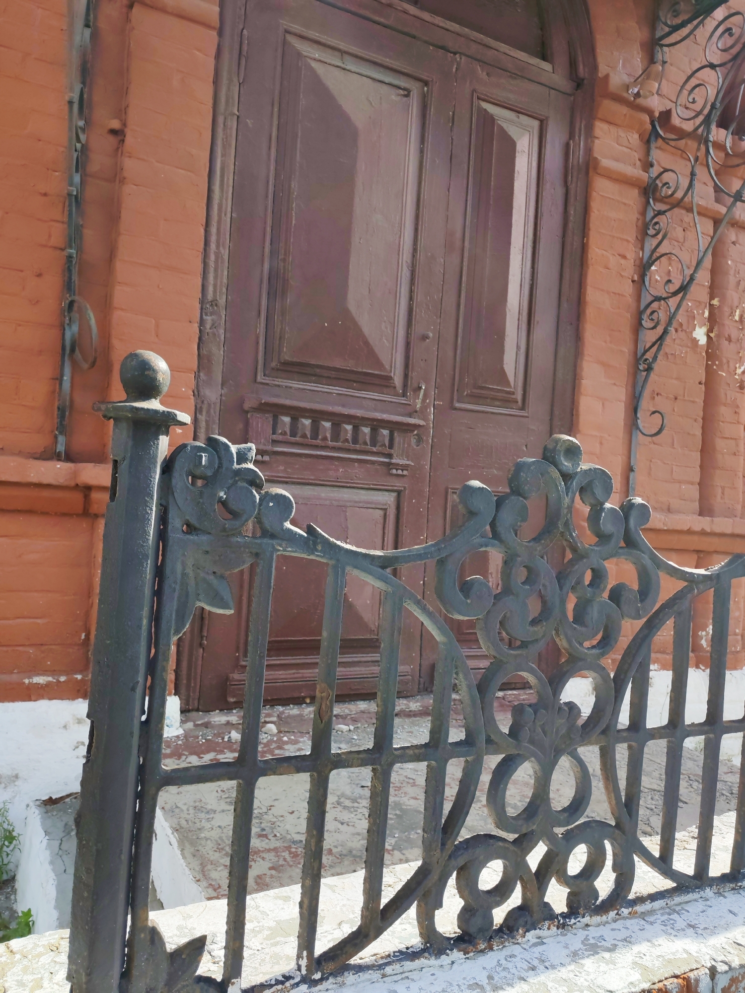 An outdoor, eye-level shot shows a close-up of a weathered, ornate black metal fence in the foreground, with an old, brown wooden door and red brick wall of a building in the background. The fence is partially obscured by a concrete base, and further details of its decorative elements, such as scrollwork and floral motifs, are visible. To the left of the fence, a black metal pole topped with a round ball marks the start of the fence. Behind the fence, a set of worn concrete stairs leads up to the wooden double doors, which are divided into panels and feature decorative molding. The brick wall exhibits signs of age, with some peeling paint visible near a wrought-iron embellishment above the door. The overall impression is one of a historic or aged urban setting.