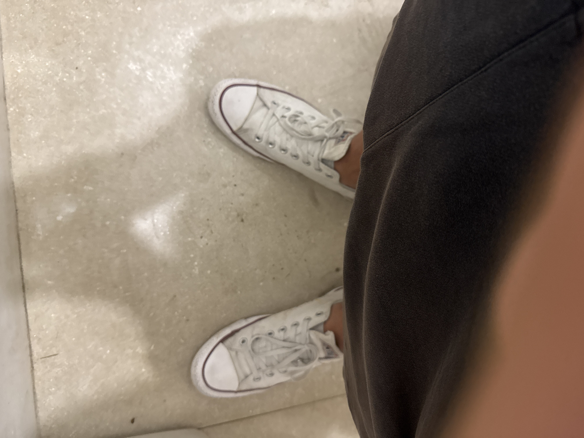 The image shows a top-down view of a person's feet wearing white Converse Chuck Taylor All-Star sneakers and dark grey pants. The shoes appear to be slightly worn. They are standing on a light-colored, speckled floor, possibly tile or terrazzo, with visible grout lines. The lighting creates a shadow on the floor behind the person's feet. The environment suggests an indoor location, possibly a restroom, hallway, or public space, based on the flooring and lighting. There are no discernable urban features or context to place the image in Kuta Selatan, Indonesia.