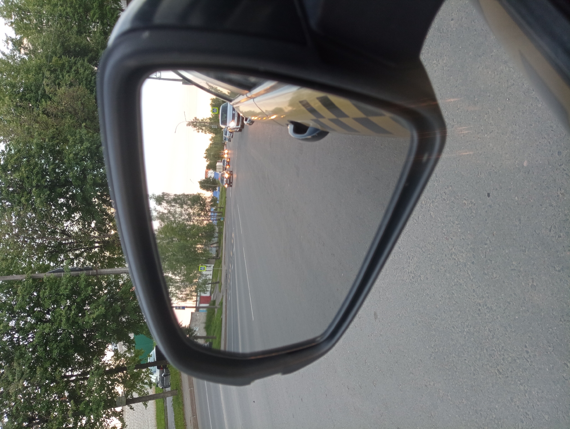 The image is a close-up, over-the-shoulder view of a car's side mirror reflecting a road with traffic. The mirror shows a street with cars, trees lining the sides, and buildings in the distance. The sky appears to be overcast, suggesting a cloudy or hazy day. The reflection in the mirror shows a road with multiple lanes and several cars. A pedestrian crossing sign is visible on the right side of the road. The car itself has a black and yellow checkered pattern on its side, possibly indicating it's a taxi. The scene appears to be in an urban or suburban setting. Without further information, it's impossible to confirm the specific location within Mahilyow, Belarus.