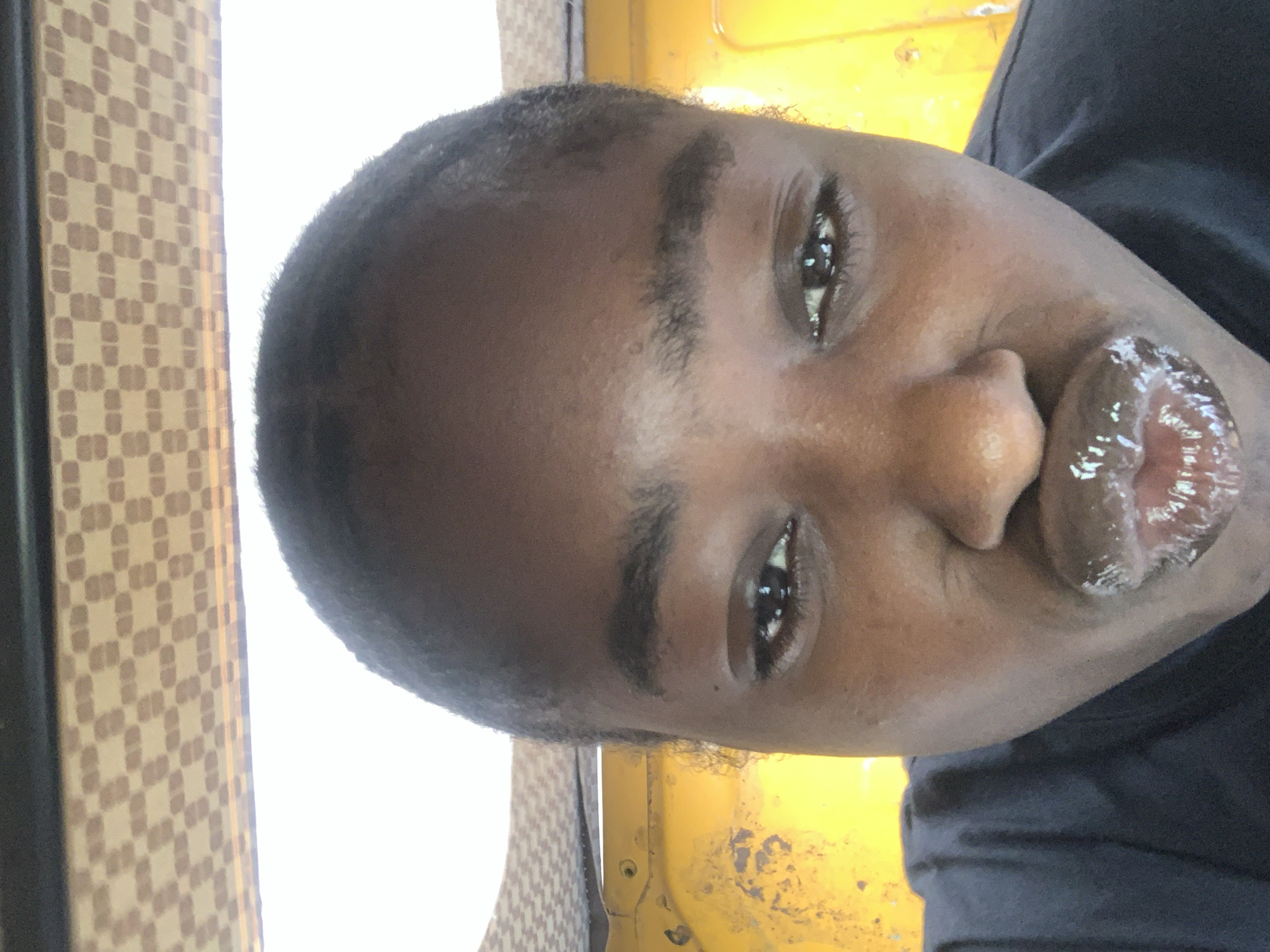 A close-up, rotated portrait of a person with dark skin and short, dark hair. They have dark eyebrows, brown eyes, a wide nose, and glossy lips. They are wearing a dark-colored shirt. The background includes a section of yellow surface and a patterned surface, likely a wall covering. The location is presumed to be within a vehicle in Rafin Par, Nigeria, based on the prompt's instructions, though this cannot be verified from the image alone.