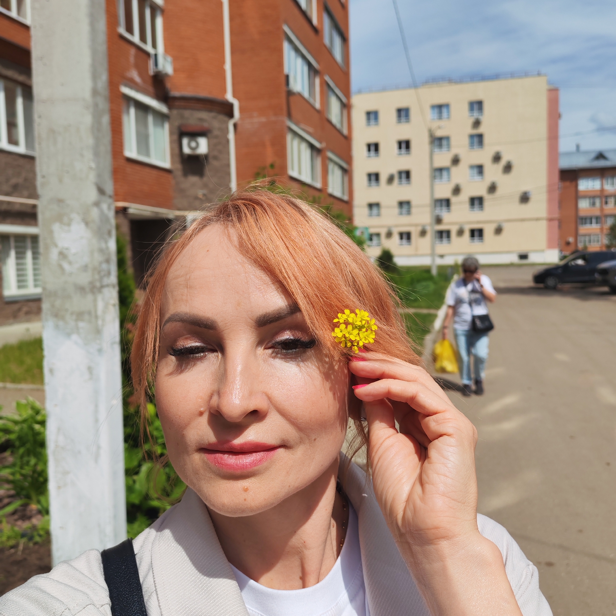 A medium close-up shot features a woman with reddish-blonde hair, light complexion, and a calm expression, possibly a selfie. She wears light makeup, a white t-shirt, and a light-colored jacket, with a black shoulder bag strap visible. A small cluster of yellow flowers is tucked into her hair above her right ear, and she appears to be holding them in place with her right hand.
The setting is an urban residential area during bright daylight, indicated by strong shadows and clear visibility. In the immediate background to the left, a tall, light-colored pole stands. Beyond it, a multi-story brick-red apartment building with white-framed windows and an air conditioning unit is visible. Further in the background, a large, light yellow/beige apartment building with numerous windows and many small air conditioning units or satellite dishes dots its facade. Another reddish building is partially seen in the far background.
A person, out of focus, walks away from the camera in the mid-ground, carrying a yellow bag. Parked cars are visible in the distance on the paved street. Green foliage can be seen on the left side of the frame, suggesting some landscaping. The sky is partially cloudy. The overall scene depicts an ordinary day in a residential neighborhood, consistent with a city like Chaikovsky, Russia. There is no visible text in the image.