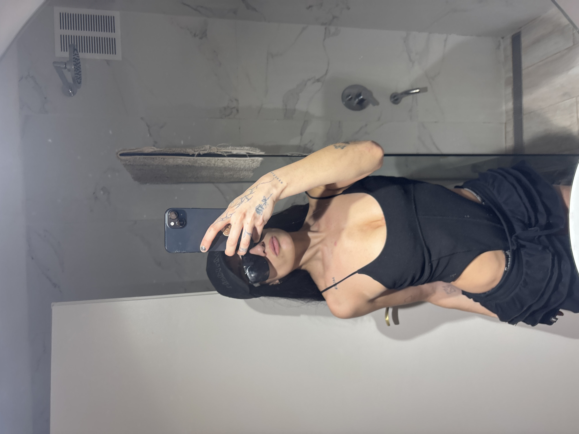 A fair-skinned person, wearing a black swimsuit-like garment and a dark baseball cap, is taking a selfie in a bathroom. They are holding a blue smartphone in their left hand. They are also wearing sunglasses.
The bathroom has white marble-patterned walls, a shower with a silver showerhead, and a silver faucet. There is a vent above the showerhead. A shelf is visible in the mirror's reflection. There are no indications that the image was taken in Buenos Aires, Argentina.