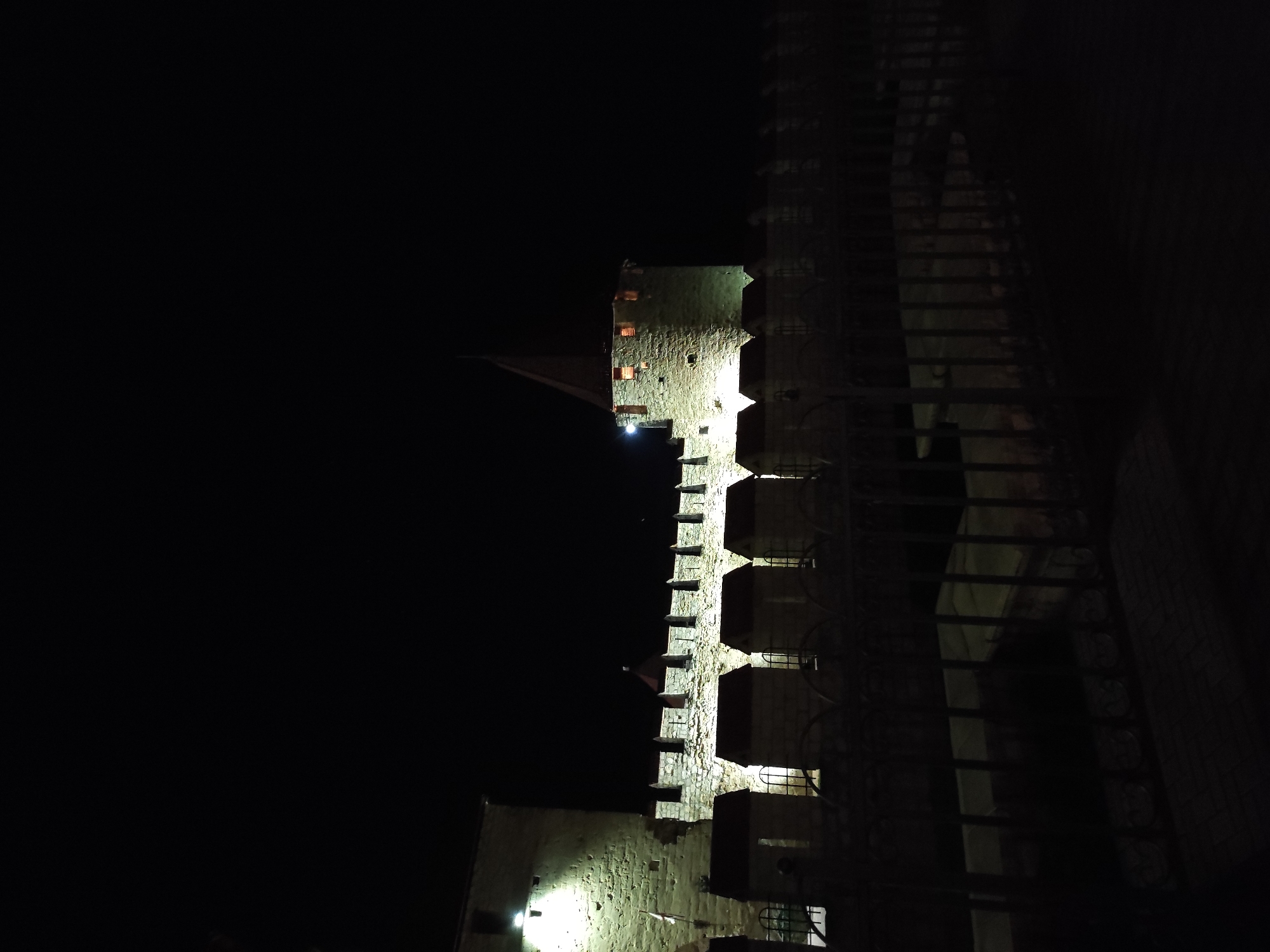 The image shows a tall stone building, likely a tower or castle, illuminated at night. The architecture is medieval in style, with crenellations and windows. The tower is lit by external sources, emphasizing its texture and form. The environment is dark, suggesting the image was taken at night in a city. A fence or railing can be seen in front of the tower. The location is in the city of Unknown, Unknown.