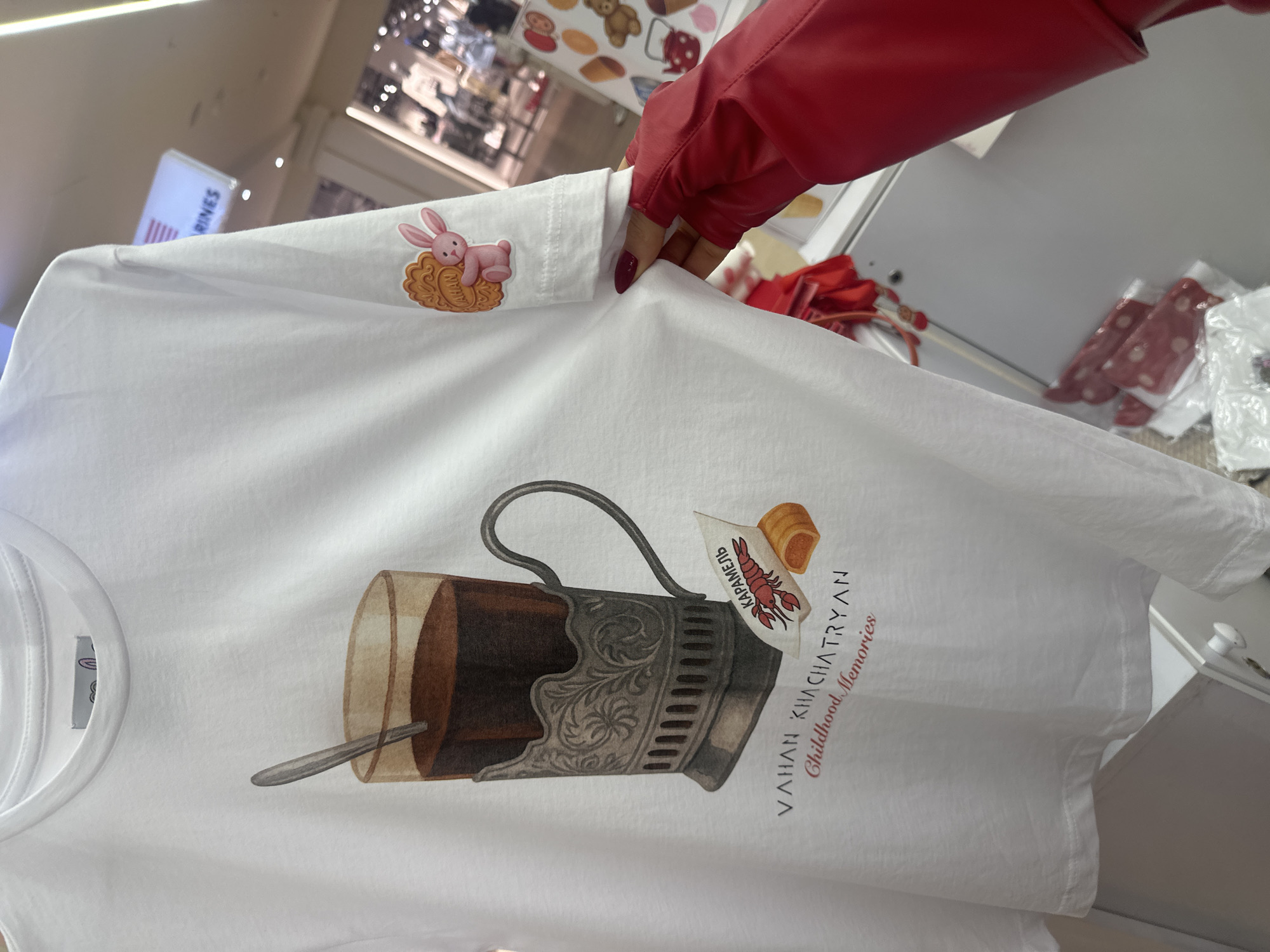 The image shows a white t-shirt being held by a person wearing a red leather jacket. The t-shirt has a graphic design featuring a traditional Russian tea glass holder with a glass of tea and a drawing of a lobster candy. Below the design, the text "VAHAN KHACHATRYAN Childhood Memories" is visible. On the sleeve of the t-shirt, there is a sticker of a pink bunny lying on a cookie. The background suggests a store environment with clothing racks and various merchandise displayed. The presence of the text "VAHAN KHACHATRYAN" hints at a connection to Yerevan, Armenia, as it could be the name of a local artist or designer.