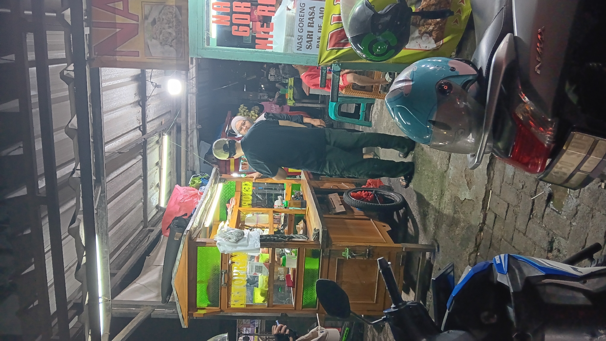 The image depicts an outdoor food stall scene at night in Depok, Indonesia. A wooden food cart with glass display shelves, illuminated by green and white lights, is the central focus. It appears to be a street food vendor selling local Indonesian dishes, as indicated by signage that reads "KETOPRAK" on the cart and other signage on a stall to the right that includes "NASI GORENG" and "MIE AYAM".
Several individuals are present. A man wearing a dark t-shirt, dark pants, and a light-colored baseball cap is standing in front of the cart, seemingly interacting with a woman. The woman is wearing a headscarf and a dark top. Another man in a red t-shirt is seated on a plastic stool to the right of the food cart. Two motorcycles are parked in the foreground, one in blue and white and another mostly visible in dark color, both with helmets resting on them. One helmet is light blue with a pink design, and another, partially visible, is green.
The environment suggests a busy street or market area with paved ground, some puddles of water, and overhead structures and lighting, indicating an urban setting at night. The overall impression is of a casual, late-night dining experience at a local street food establishment.
