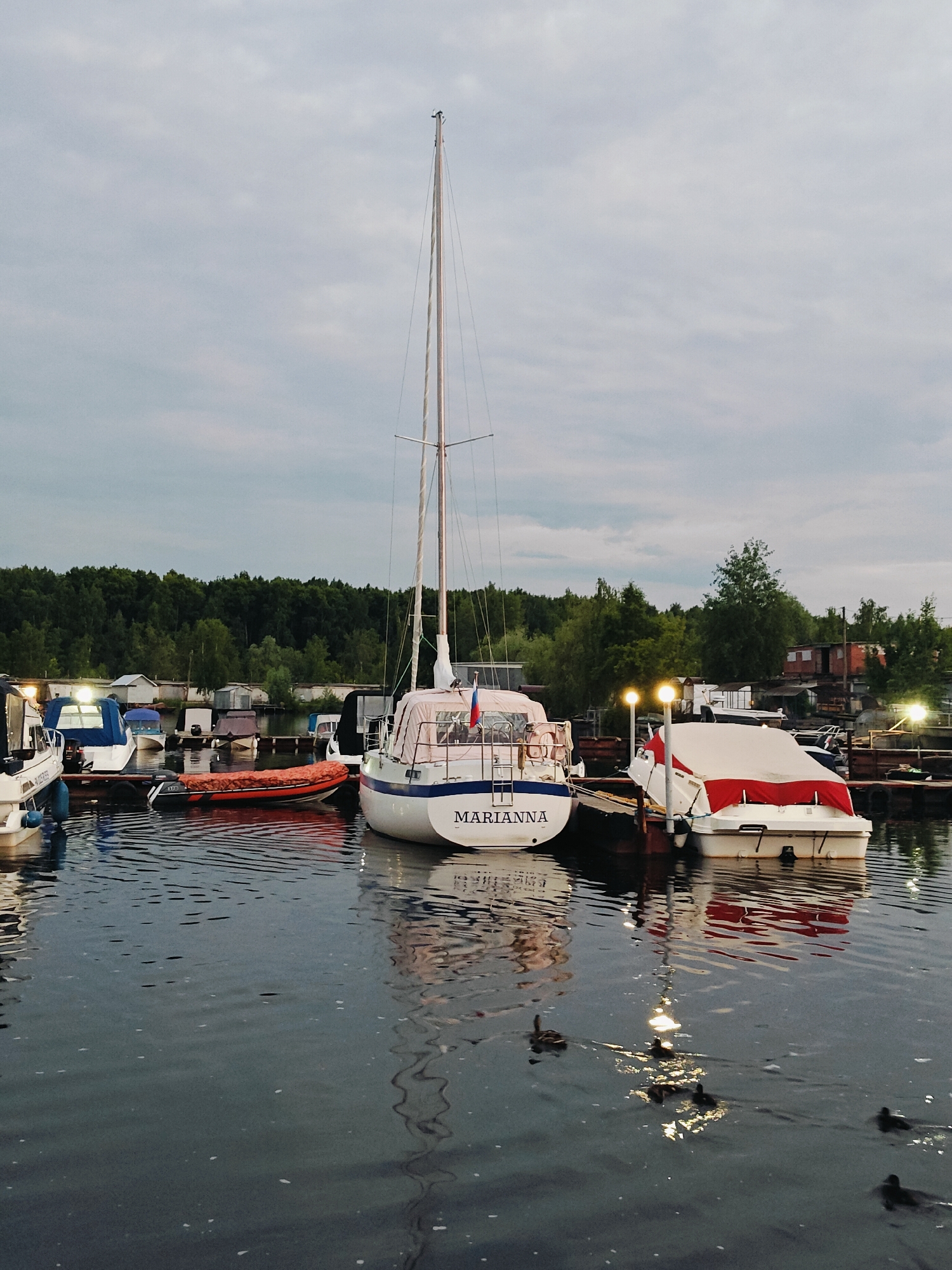 In the city of Chaikovsky, Russia, a serene evening scene unfolds at a marina. The water is calm, with gentle ripples reflecting the overcast sky and the lights of the docked boats. Several boats are moored, including a prominent white sailboat named "MARIANNA," its mast reaching towards the sky. Adjacent to it, another white boat is covered with a red and white tarp. A smaller orange inflatable boat is also visible. In the background, a lush green forest borders the marina, and a line of small buildings stands along the shore. The sky is a muted gray, suggesting dusk or an overcast day. Several ducks are swimming in the water, adding a touch of natural life to the otherwise still environment.