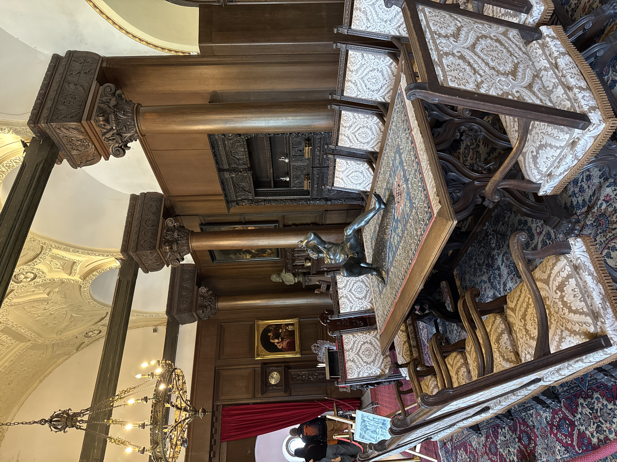 This image depicts the interior of what appears to be the dining hall of the Reichsburg Cochem castle, in Cochem, Germany. The room is characterized by dark wood paneling, ornate columns, and a detailed chandelier. A large, rectangular table with a patterned tablecloth is surrounded by chairs with patterned upholstery. A bronze statue stands on the table. Artworks, including paintings and sculptures, adorn the walls, and a clock is visible on the wall. At the far end of the room, red curtains drape a doorway, through which two individuals are visible. The floor is covered with a patterned rug.