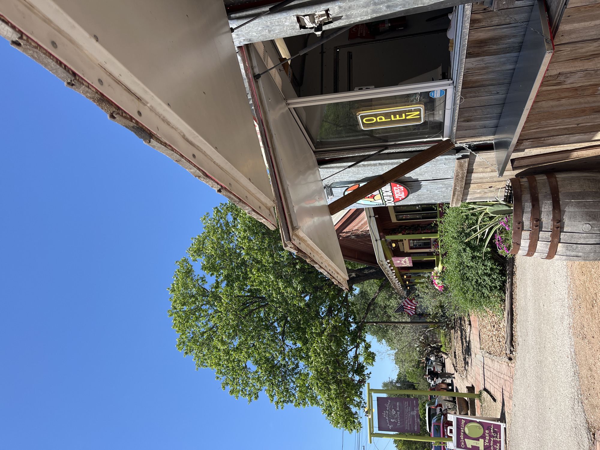 The image captures a street scene, possibly in New Braunfels, Texas. An "Open" sign is visible on a building with a service window or awning. Signs indicate nearby establishments: "ROCKY MOUNTAIN CREPES," and "The Green Olive Tasting Room", and another celebrating "10 Years". There are trees along the street, and a street lined with businesses is visible in the distance. The sky is clear and blue. A barrel rests beside the building.