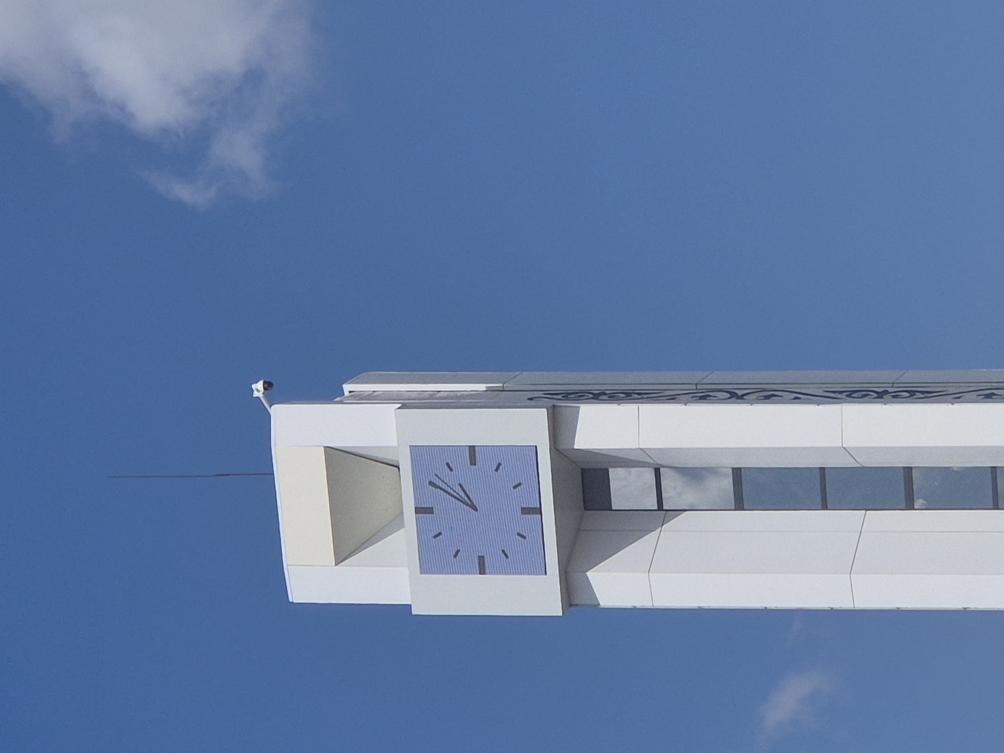 A close-up, low-angle shot captures the top section of a modern, white clock tower against a clear blue sky. The tower features a large, square clock face with a light blue background and black hour markers. The clock hands are pointing to approximately 1:05. At the very top of the tower, a small, white security camera is mounted on a horizontal arm. A thin, black antenna or lightning rod extends vertically from the apex of the tower. The clock tower is located in the city of Kokshetau, Kazakhstan.