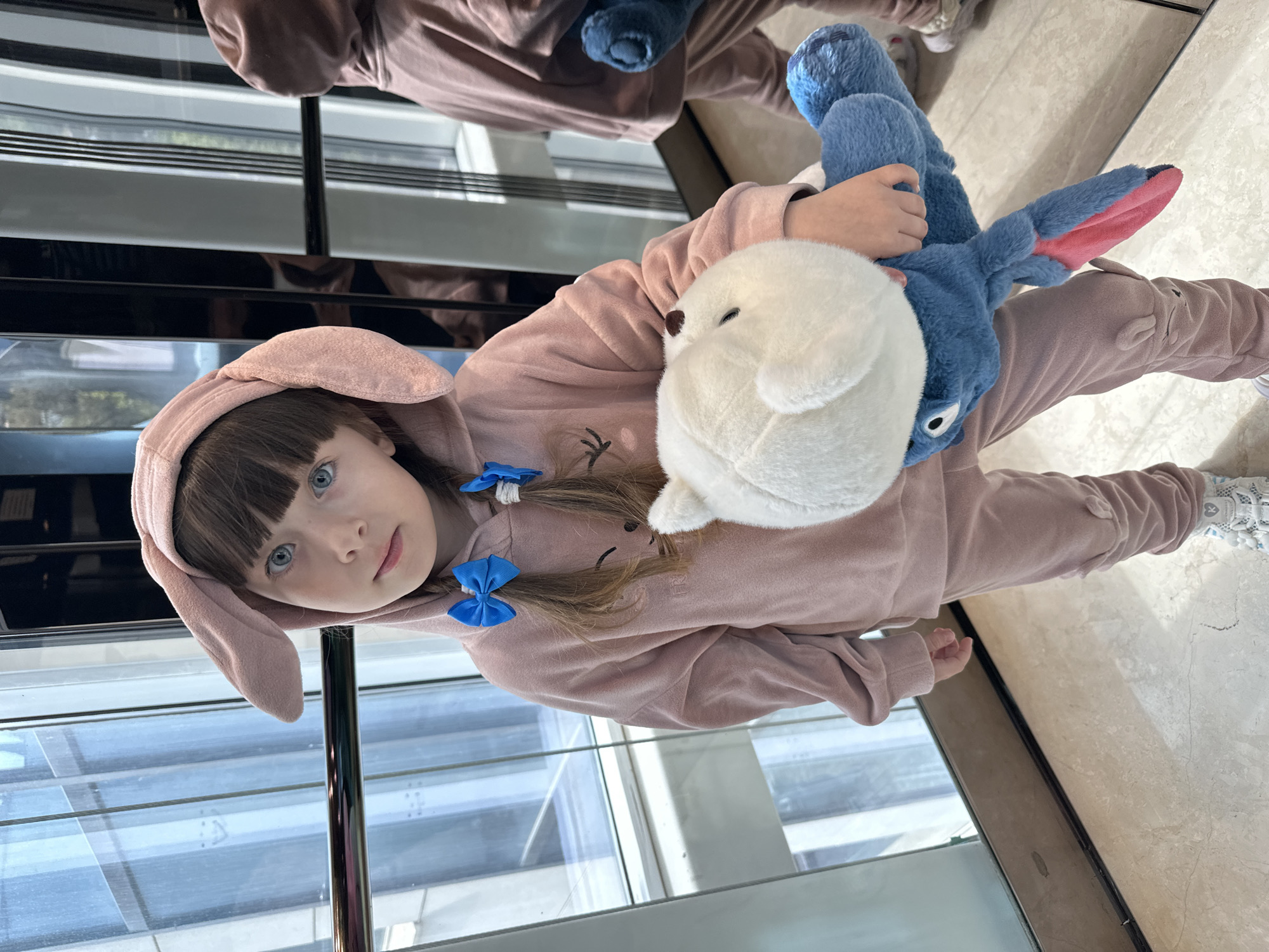 A young girl, approximately 5 to 7 years old, is the central subject of the image, captured indoors in what appears to be a modern building in Sochi, Russia. She has bright blue eyes and dark brown hair with bangs, adorned with two small blue bow hair ties.
She is dressed in a soft, pink, plush onesie or hooded jumpsuit, complete with bunny ears on the hood and subtle embroidered details resembling a face on the chest. The girl is holding two stuffed animals: a fluffy white one, possibly a polar bear cub, and a larger blue character that resembles Stitch from Disney's Lilo & Stitch. She looks directly at the camera with a calm and engaging expression.
The background consists of large, reflective glass panels or windows, possibly featuring horizontal blinds or structural elements, which reflect parts of the room and an indistinct outdoor scene. The floor is a light-colored, highly reflective surface, likely polished marble or tile. A dark, reflective vertical column or frame is also visible. The girl's own reflection can be seen in the background's glossy surfaces. The lighting suggests it is daytime. No visible text is present in the image.