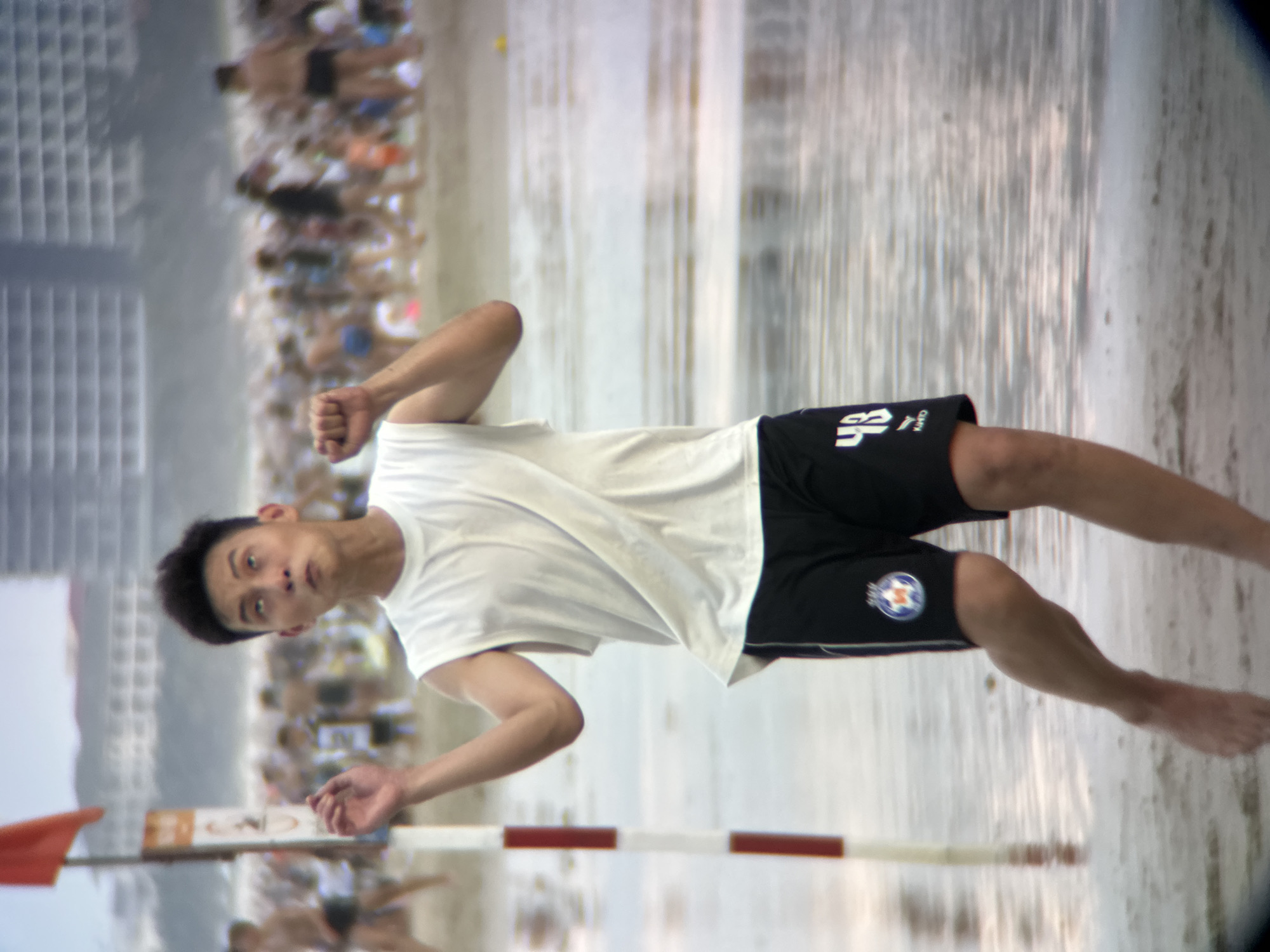 A young Asian man in a white sleeveless shirt and black shorts with white trim and the number 43 on his right leg is standing on a wet beach, looking up with his mouth open. He has dark hair and is mid-motion, with his left arm raised and his right fist clenched. In the background, a crowd of people are visible on the beach, and buildings with grid-like windows rise in the distance. A red flag flies on a pole to the left of the man. The scene appears to be on a beach in Đà Nẵng, Vietnam.
