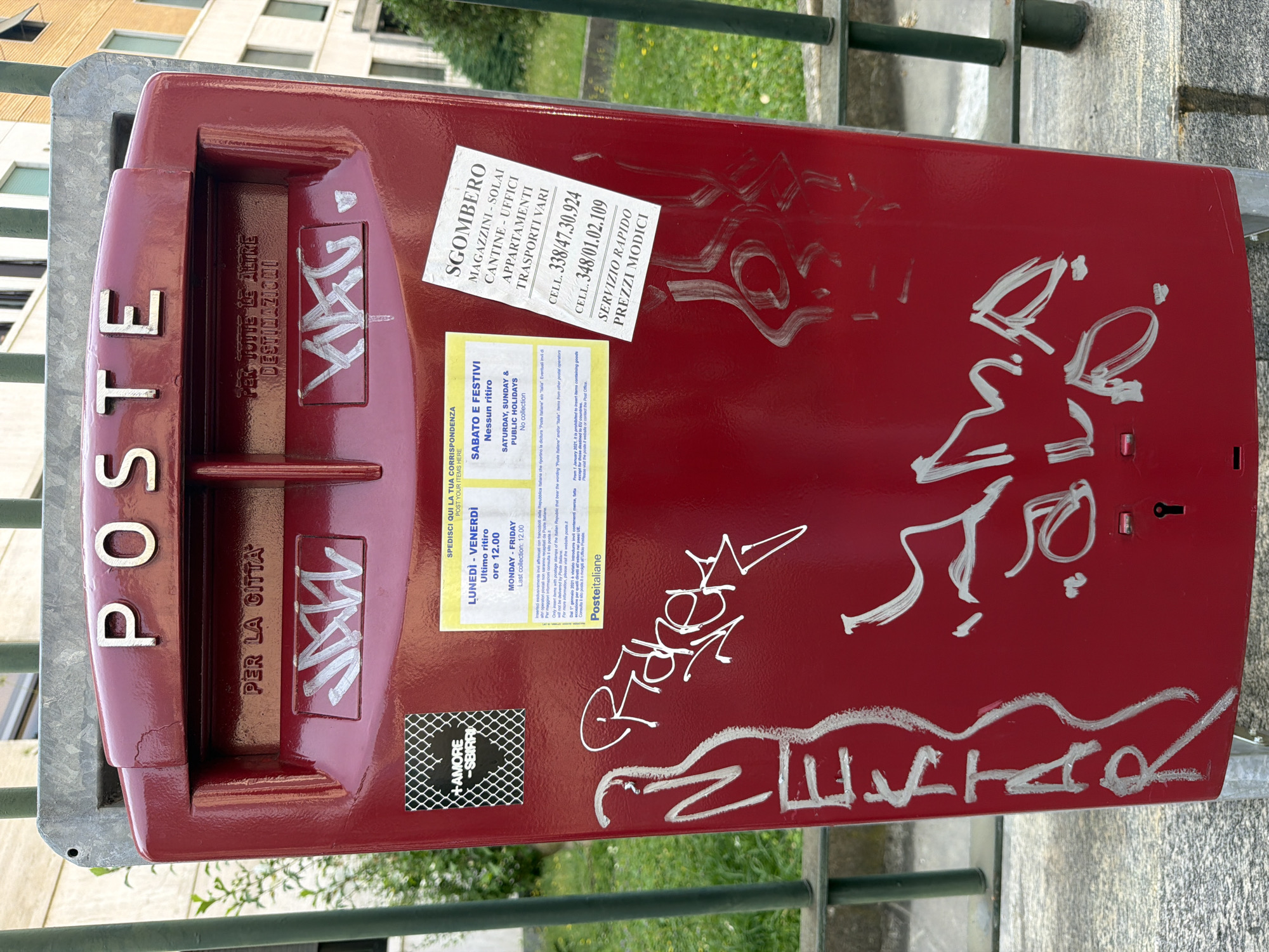 An outdoor, eye-level, medium shot shows a close-up of a red, public postal mailbox. The mailbox has the word "POSTE" embossed in silver capital letters across the top. Below that, two slots are labeled "PER LA CITTA" and "PER TUTTE LE ALTRE DESTINAZIONI." The mailbox is covered in white graffiti, including names and abstract shapes. Attached to the mailbox are several stickers. On the left, a black and white sticker has "+AMORE SBIRRI" written on it. To the right, a yellow sticker provides information about collection times. A white sticker with black text lists services and phone numbers, including "SGOMBERO MAGAZZINI - SOLAI CANTINE UFFICI APPARTAMENTI TRASPORTI VARI" and phone numbers starting with "338" and "348." The mailbox is mounted on a grey metal frame, and behind it, a blurry green leafy plant and a portion of a building are visible. The ground appears to be a paved surface.