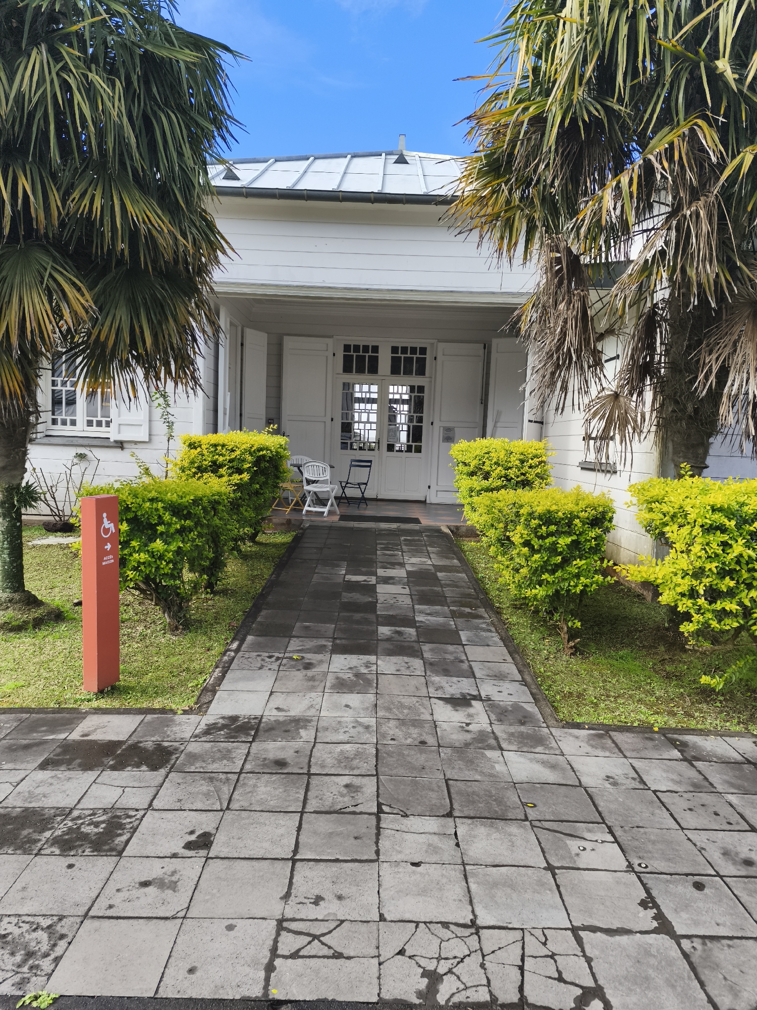 The image shows the entrance to a white building, possibly a public facility or residence, with a walkway leading up to it. Two palm trees frame the entrance. The walkway is paved with square grey tiles, some of which are wet and appear stained. On the left side of the walkway, there is a red post with a white accessibility symbol and text that reads "ACCES MAISON." Adjacent to the post are neatly trimmed green bushes and a patch of grass. The building itself has white clapboard siding, white double doors with glass panes in the center, and white shutters on the windows. A white metal roof with visible seams covers the structure. The sky above is a clear blue. The overall scene suggests a well-maintained entrance to a building in a tropical or subtropical climate. There are no people visible in the image.