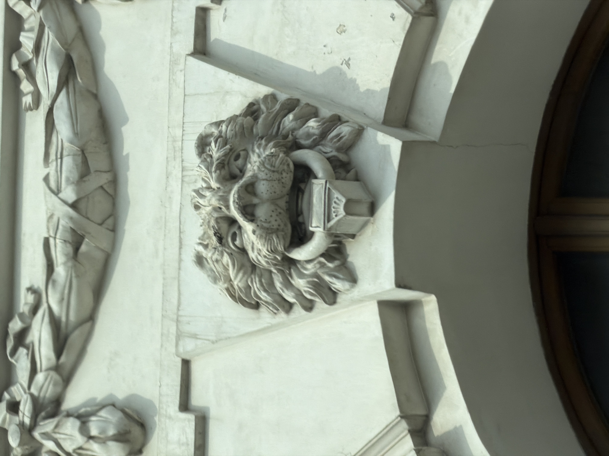 The image depicts a detail of an architectural facade, likely in the city of Unknown, Unknown. A sculpted lion's head, rendered in a pale material such as stone or plaster, is prominently featured. The lion's head serves as a decorative element, possibly part of a door or window surround, and includes a metal ring held in its mouth. To the left of the lion's head, there is another sculptural element, appearing to be a draped fabric or garment, also rendered in the same pale material. A portion of a window frame is visible on the right side of the image. The architectural style suggests a classical or neoclassical influence. The facade is lit by natural light, creating shadows that accentuate the sculpted details. The overall impression is one of ornate and historical architecture.