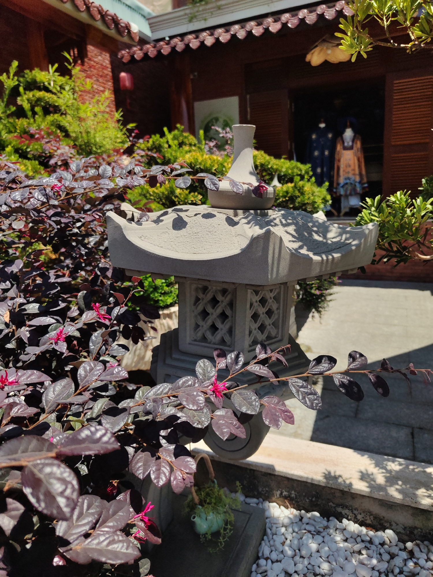 The image shows a Japanese-style stone lantern in a garden setting. The lantern is gray, with a multi-tiered roof and lattice-like openings. A plant with dark purple leaves and small pink flowers is prominent in the foreground, partially obscuring the lantern. Behind the lantern, there's a building with a traditional Vietnamese or Chinese architectural style. In the background, inside the building, two figures can be seen wearing what appears to be traditional Vietnamese clothing (likely Ao Dai). The ground is paved, and white pebbles are visible in a recessed area around the lantern. The location is likely in a garden or courtyard in Cam Ly - Đà Lạt, Vietnam, given the architecture and style of the scene.