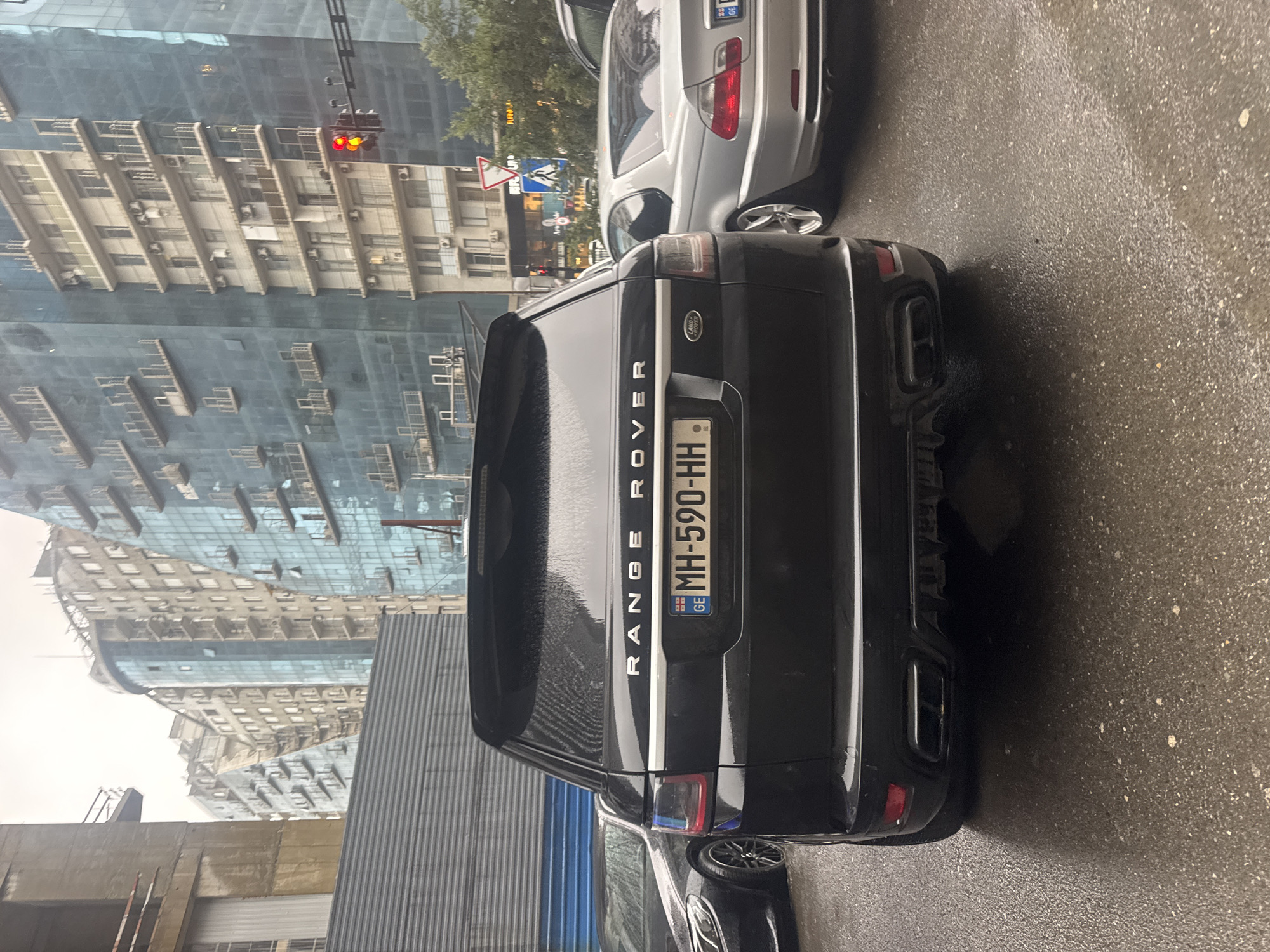 The photograph depicts the rear of a black Range Rover with Georgian license plate MH-590-HH. The vehicle is parked on a wet, paved surface, suggesting recent or ongoing rain. To the right of the Range Rover, a silver BMW is partially visible. In the background, there are modern, high-rise buildings with glass facades. Traffic lights and street signs are also visible, indicating an urban setting. The architecture and signage suggest the location is in Tbilisi, Georgia.