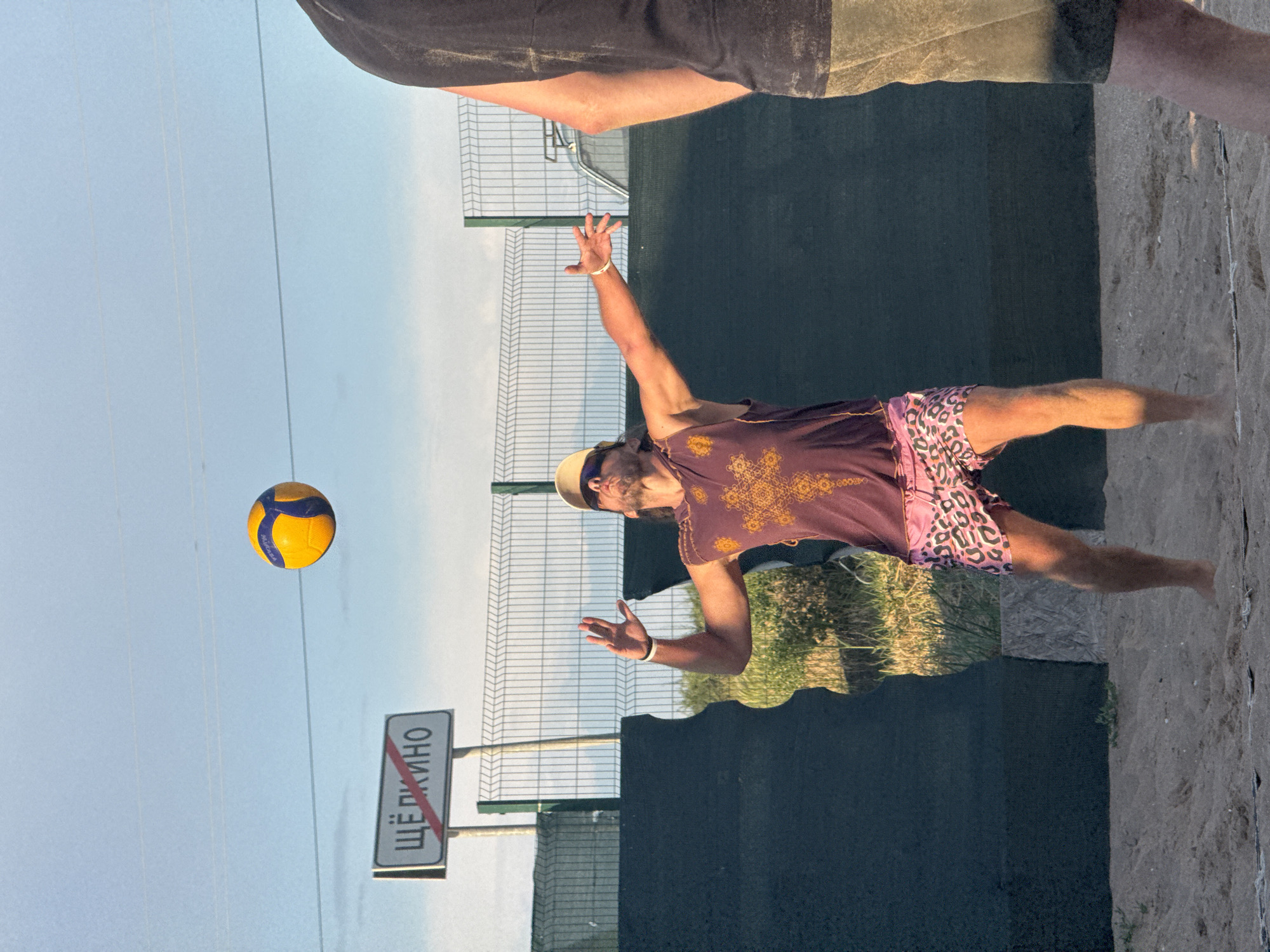 A man in a purple tank top with a gold star pattern and pink leopard-print shorts is playing beach volleyball. He is in mid-air, reaching up to hit the ball, which is a yellow and blue Mikasa brand volleyball. The man is wearing a light-colored baseball cap and sunglasses. In the background, a sign with Russian lettering, "ЩЁЛКИНО", indicates the location is the urban settlement of Shchyolkino. A fence and dark green netting are also visible behind the man. Another person's legs are visible in the foreground on the right side of the image, suggesting a game is in progress. The setting is a sandy beach volleyball court under a clear blue sky with some power lines stretching across it.