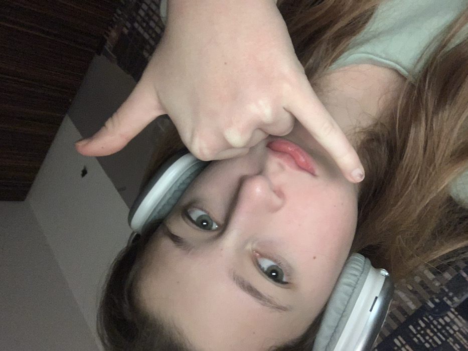 A fair-skinned young woman with hazel eyes and light brown hair is pictured in a selfie-style, upside-down shot. She's wearing grey and white headphones. One hand is near her face, gesturing with the index and pinky finger extended. Her expression is somewhat neutral, but her lips are slightly parted. A dark wooden surface is visible in the background.