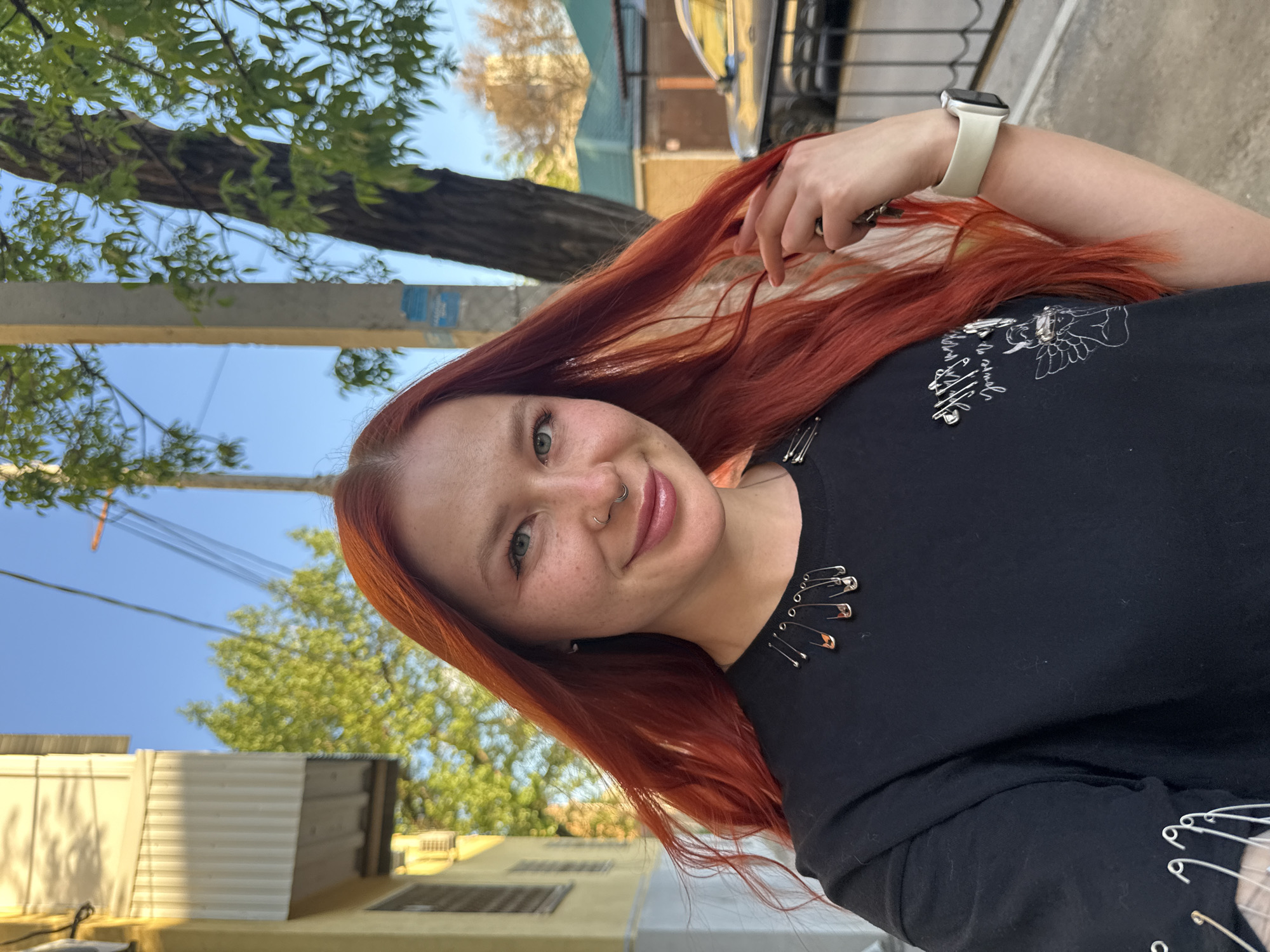 A young woman with vibrant red hair and freckles is posing outdoors, presumably in Rostov-on-Don, Russia. She is wearing a black t-shirt adorned with safety pins, and a white smartwatch is visible on her wrist. The background consists of urban elements such as buildings, trees, and power lines against a clear blue sky. The lighting suggests it is daytime.