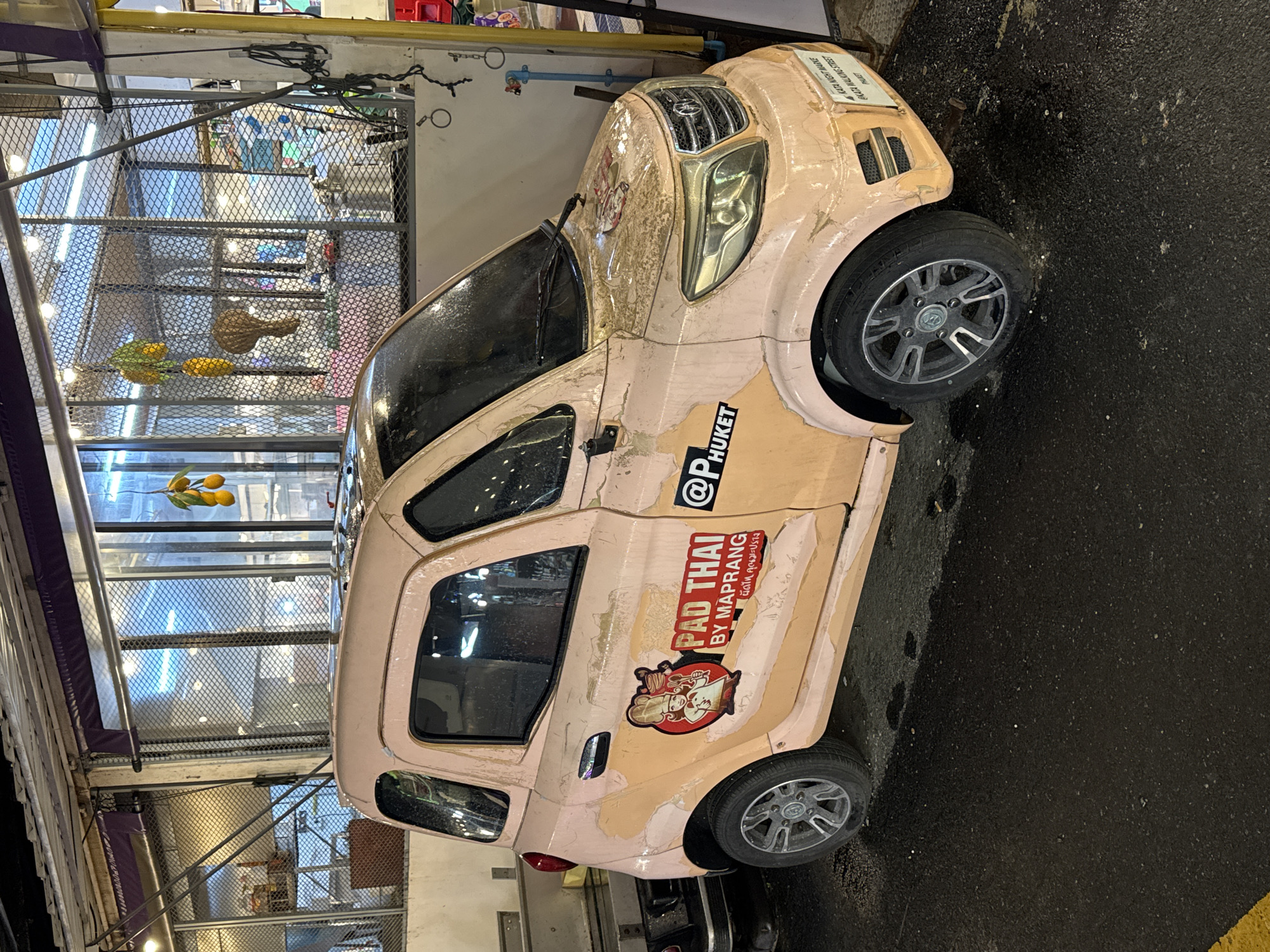 A light pink, small, four-wheeled vehicle, possibly a car or a small van, is parked on an asphalt surface. The vehicle has a faded paint job with areas of peeling. On its side, there are two stickers. The first sticker, located on the rear passenger door, features a cartoon chef with a hat and apron, holding a wok, with the text "PAD THAI BY MAPRANG" above it. Below this text are Thai characters. The second sticker, placed on the front passenger door, displays "@PHUKET" in black letters. A white license plate with black text reading "KATA NIGHT MARKET @KATA WALKING STREET PHUKET" is affixed to the front bumper. The vehicle is partially obscured by a dark object on its left. In the background, a building with a metal mesh facade is visible, with yellow hanging decorations resembling fruit. The ground is wet, with some small white specks scattered on it. The overall scene appears to be an outdoor marketplace.