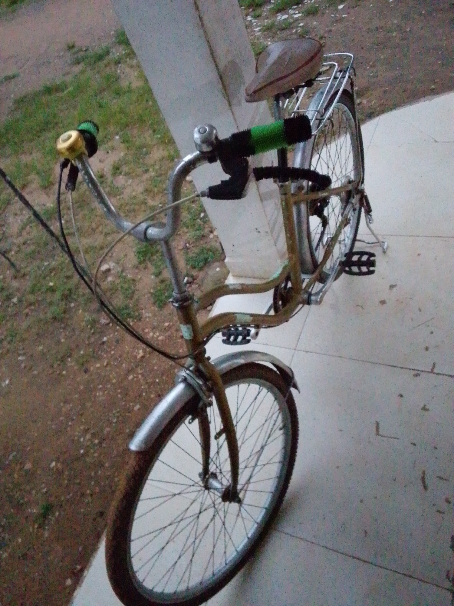 The image shows a stationary, vintage bicycle parked on a tiled surface, likely outdoors, next to a concrete pillar and dirt ground with sparse vegetation. The bicycle is a light brown color and appears to be in a state of moderate disrepair, with some rust visible on the frame. It features a brown saddle, a rear rack, and two pedals. The front handlebar has a golden bell and a green grip on the left side, and a silver bell on the right. The bicycle is positioned to face towards the left of the frame, with its front wheel prominently in the foreground and its rear wheel visible further back. The lighting suggests it is daytime, possibly overcast. The location is identified as Serti, Nigeria.