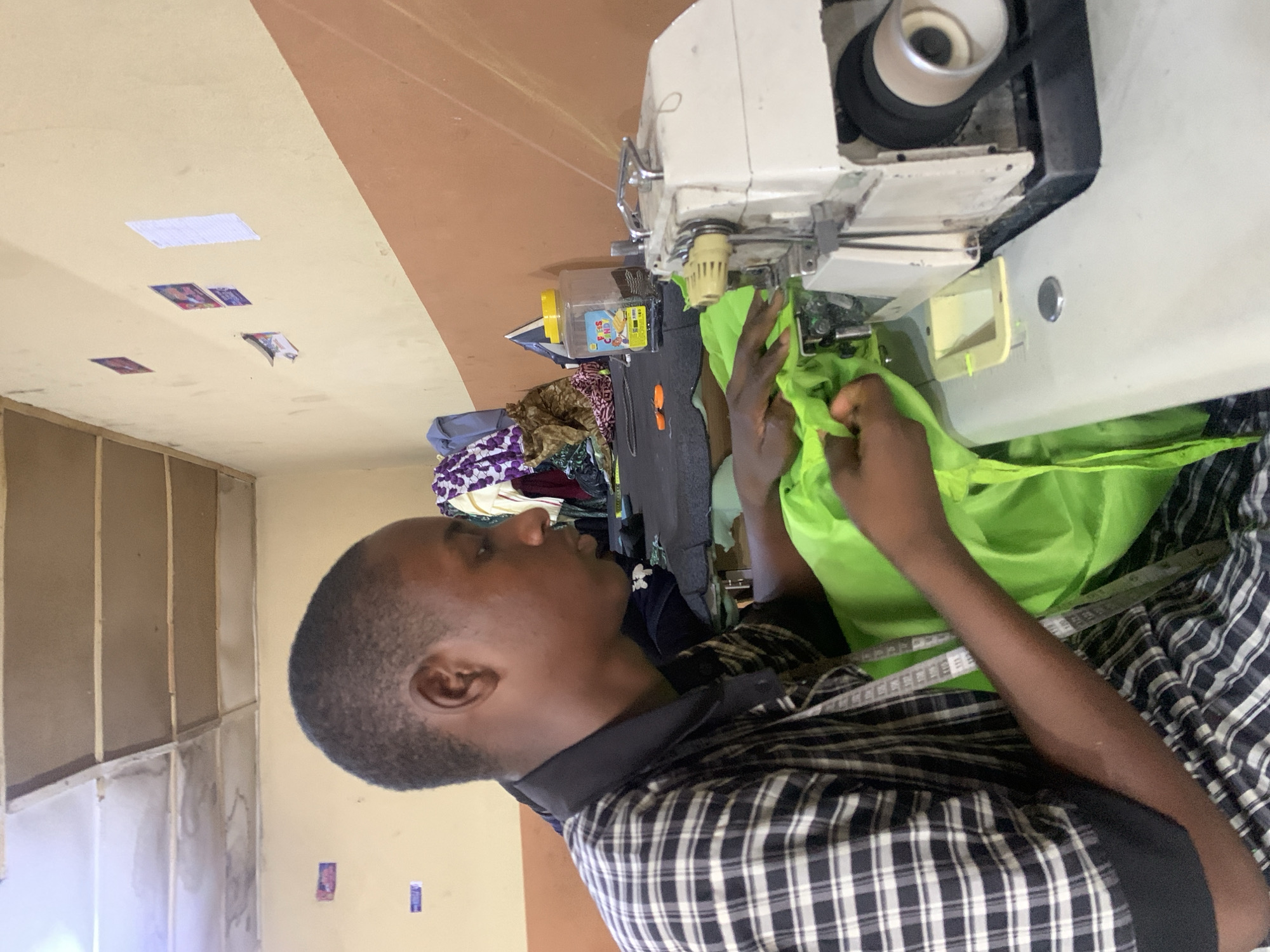 A person is positioned at a sewing machine in a room with a tan-colored wall, located in Jos, Nigeria. The individual is wearing a checkered shirt and a measuring tape around their neck, with their hands holding a bright green fabric as they operate the sewing machine. Assorted fabrics and sewing accessories are visible on the workspace around the machine. Papers are also visible on the walls.