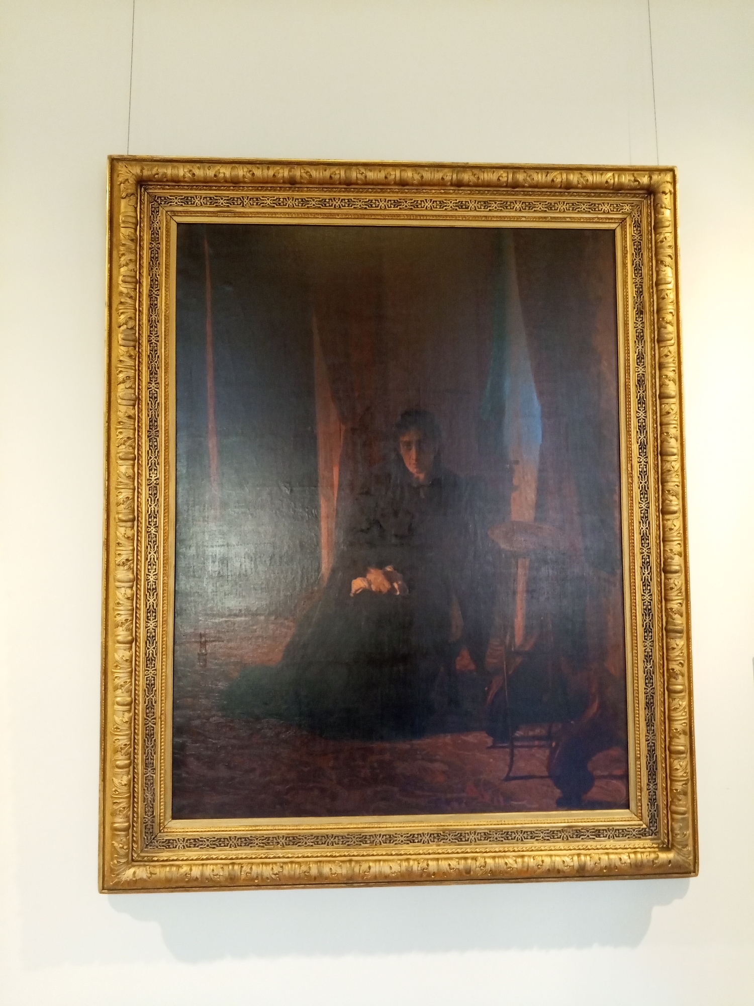 The image shows a painting displayed on a white wall. The painting is in a large, ornate gold frame. The subject of the painting appears to be a portrait of a woman in a dark dress, seated in a dark room. The painting is hanging by a thin wire from the ceiling. This painting is located in Saint Petersburg, Russia.