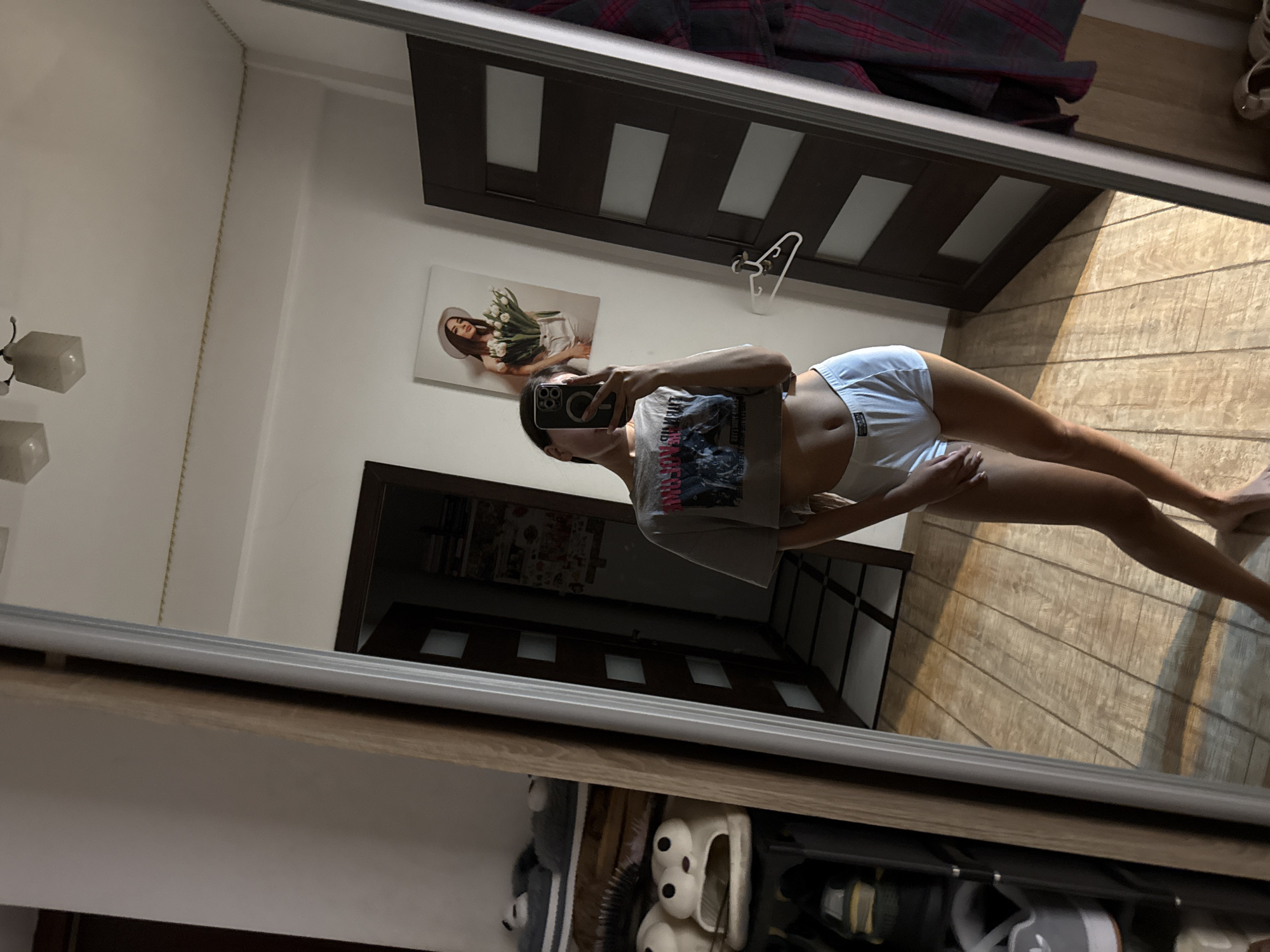 A full-body mirror selfie is taken by a young woman wearing a gray crop top and white shorts. She is standing in what appears to be a bedroom with a wooden floor and a large mirror. To her left, there is a wardrobe with shelves holding various items, including slippers. To her right, there is a dark brown door with rectangular panels. A framed picture of a woman with flowers is hanging on the wall above the door. The lighting suggests it is daytime, with sunlight streaming in and casting shadows on the floor. There is no information available to determine the location of this image in Chișinău, Moldova.