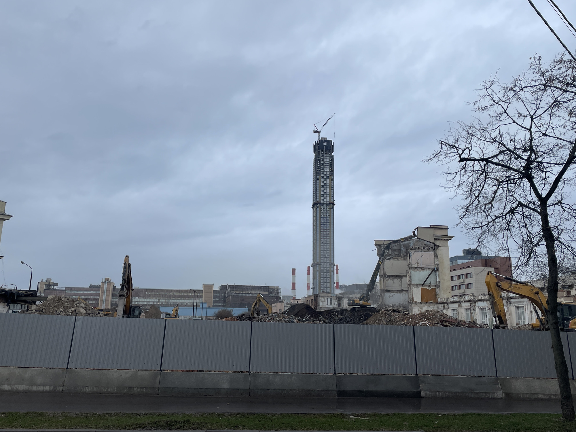 The image depicts an urban demolition site in Moscow, Russia. A grey corrugated metal fence runs along the foreground, separating the viewer from the construction zone. Behind the fence, there is rubble and several yellow excavators actively demolishing what appears to be an old building. A skyscraper under construction with a crane at its apex dominates the skyline. In the background, there are several industrial buildings and red and white striped smokestacks. A bare tree stands to the right of the frame. The sky is overcast.