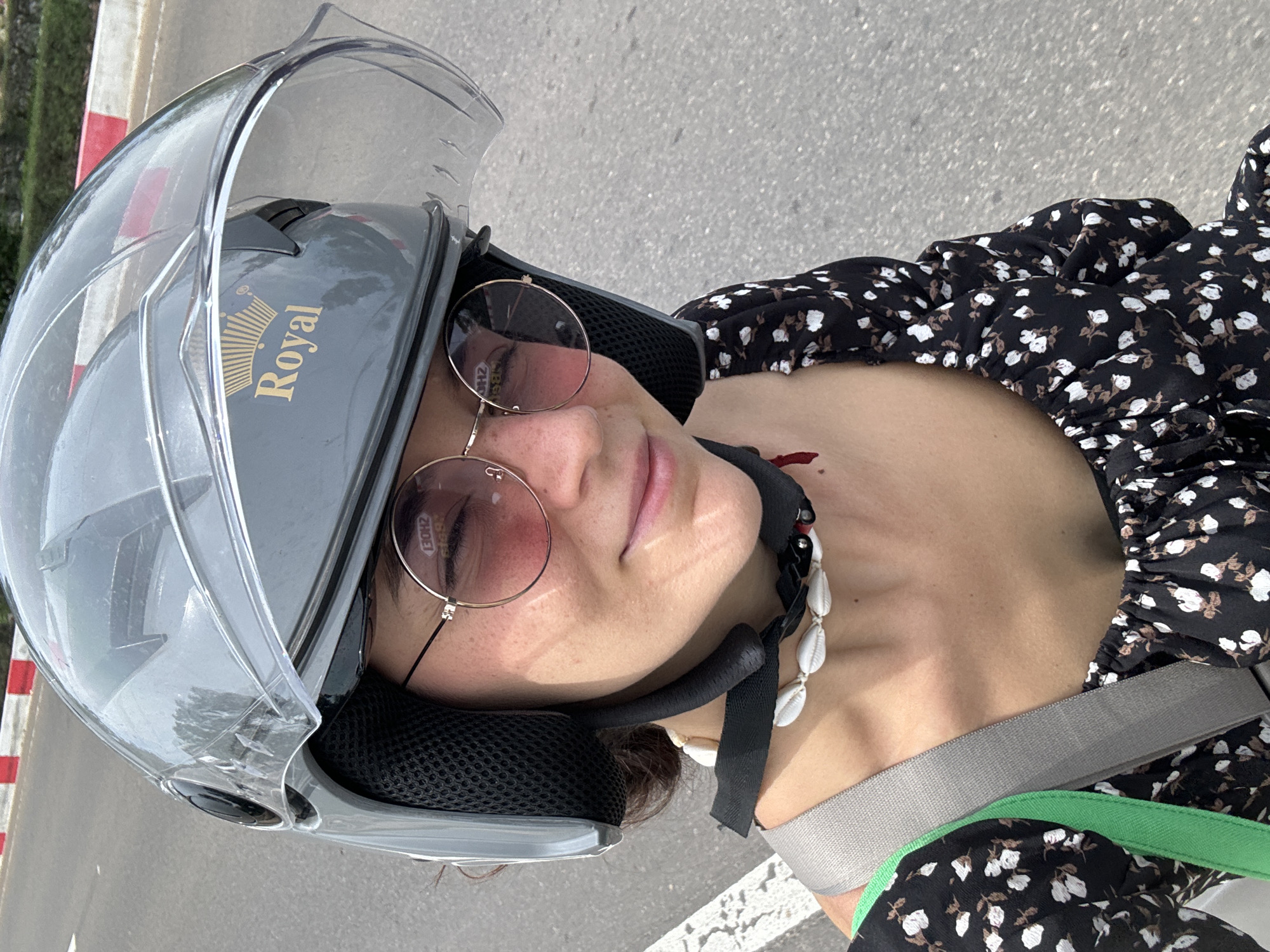 An outdoor, close-up, and vertically oriented shot shows a person, likely female, from the chest up, looking towards the camera. The person is wearing a grey motorcycle helmet with the clear visor lifted, revealing round, rose-tinted sunglasses. A slight, relaxed smile is visible on their face.
They are dressed in a black top or dress adorned with a white and brown floral pattern. A distinctive necklace made of white cowrie shells is worn around their neck. A grey strap, possibly from a bag or a seatbelt, crosses diagonally over their chest.
The background consists of an asphalt surface, indicative of a road or paved area, with a white road marking visible. To the left, a red and white striped barrier or pole can be seen. The lighting suggests bright daylight, likely a sunny day.
Visible text on the helmet includes the brand name "Royal" with a crown logo. A reflection in the sunglasses shows faint text, possibly "130HZ". The scene conveys a relaxed moment, potentially during travel on a scooter or motorcycle, in a location like Mũi Né, Vietnam.