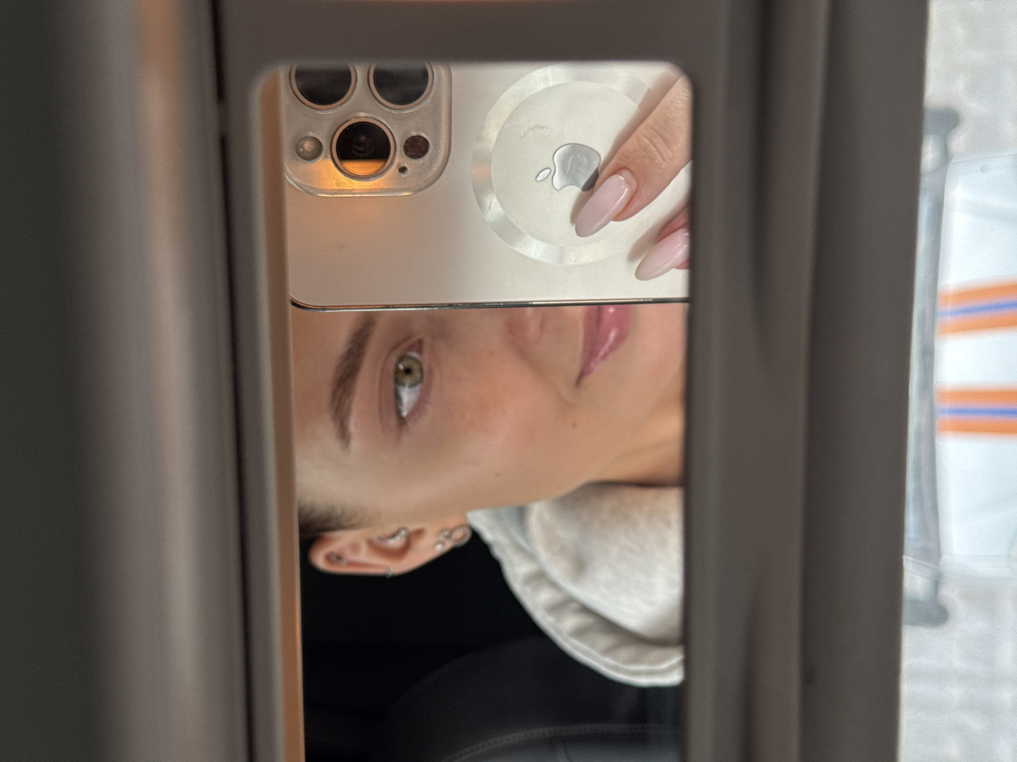 The image displays a mirror selfie, captured with a smartphone. The main subject is a person, likely a young woman, whose face is visible from the left eye to the lower ear and part of the mouth. She has green eyes and well-defined eyebrows. Her left ear features multiple piercings, including helix, conch, and lobe piercings. She appears to be wearing a light-colored hooded sweatshirt or top.
The selfie is taken using the back camera of a smartphone, which is clearly reflected in the mirror. The phone appears to be an iPhone, recognizable by its distinct camera module with three lenses and a flash/LiDAR sensor. An Apple AirTag, featuring the iconic Apple logo, is visibly attached to the back of the phone, possibly integrated into its case. A hand with neatly manicured, almond-shaped nails, painted a light pink or nude color, is seen holding the phone, with a finger gently resting on the AirTag.
The image is framed by dark, vertical borders on the left and right, suggesting it's taken through a mirror, possibly a car's rearview mirror or a compact mirror. The person's head is tilted sideways to the right, indicating the orientation of the mirror or how the photo was taken. In the blurred background, beyond the mirror's frame, faint horizontal orange and blue stripes are discernible, but the specific location cues for Chaikovsky, Russia, are not visible in the immediate reflection. The lighting is soft and consistent, highlighting the details of the phone and the person's features.