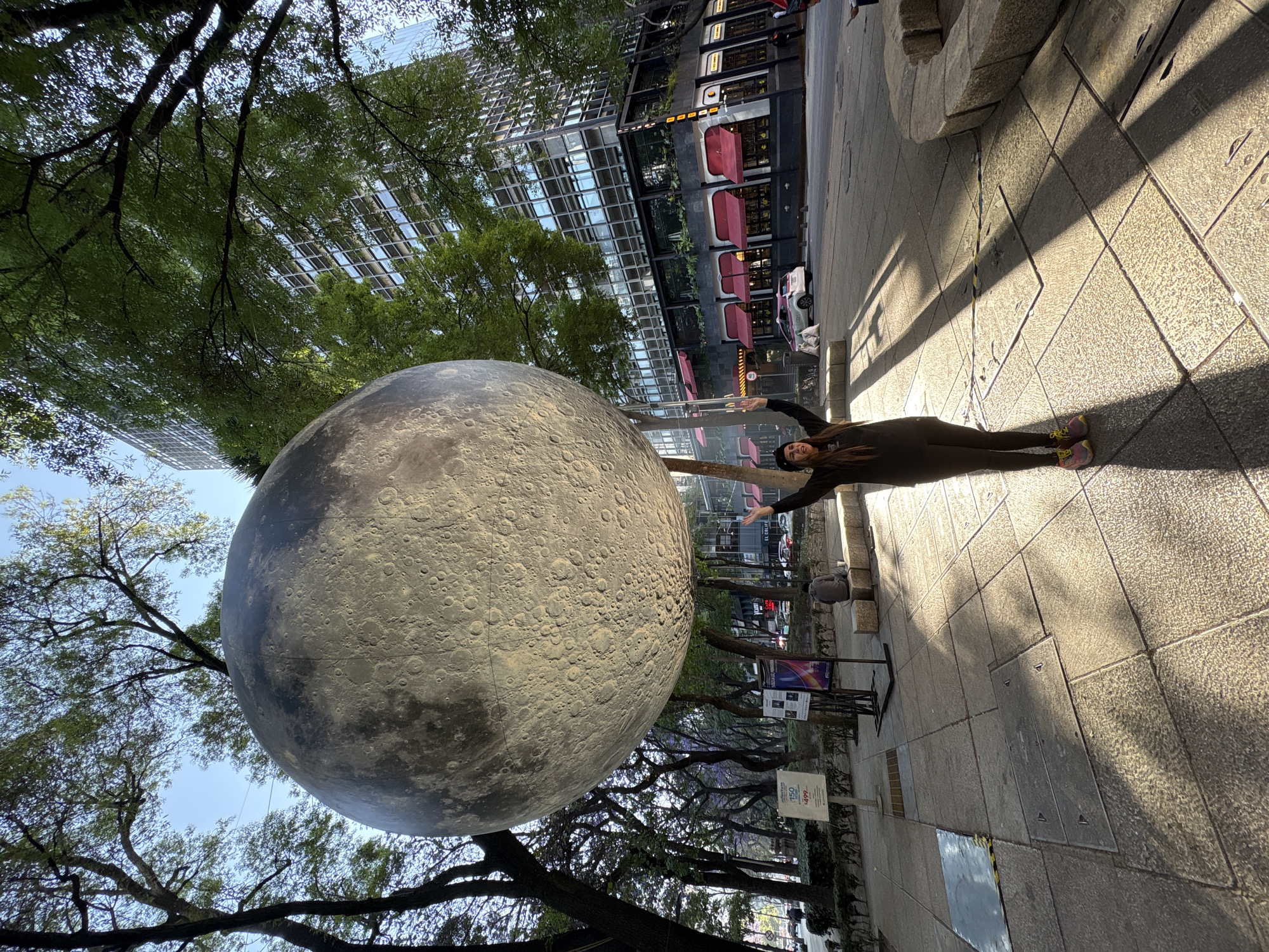 An outdoor urban scene is depicted on Paseo de la Reforma in Mexico City, Mexico, adjacent to the Torre Prisma building and Cantina Riviera del Sur restaurant. A large, spherical sculpture, textured to represent the Moon's surface with craters, is suspended centrally within the frame. A female individual stands directly below the sculpture, facing the camera, with both arms raised overhead, palms open, creating the optical illusion of supporting it. She wears dark attire and multi-colored athletic shoes. To her left, in the mid-ground, a male individual is seated on a stone bench, facing away. The foreground comprises a textured concrete sidewalk with square tiles and metal utility covers. Overhead, a canopy of green-leaved trees is visible against a clear sky. To the right, a tall, multi-story building with a glass and concrete facade (Torre Prisma) dominates, its ground floor occupied by a commercial establishment with red awnings and large windows (Cantina Riviera del Sur). A paved street, containing parked and moving vehicles including a white and pink taxi, runs between the sidewalk and the building. Exhibition stands and a sign advertising 'Finfun' are present on the left sidewalk. The scene is brightly lit by natural sunlight, casting prominent shadows.