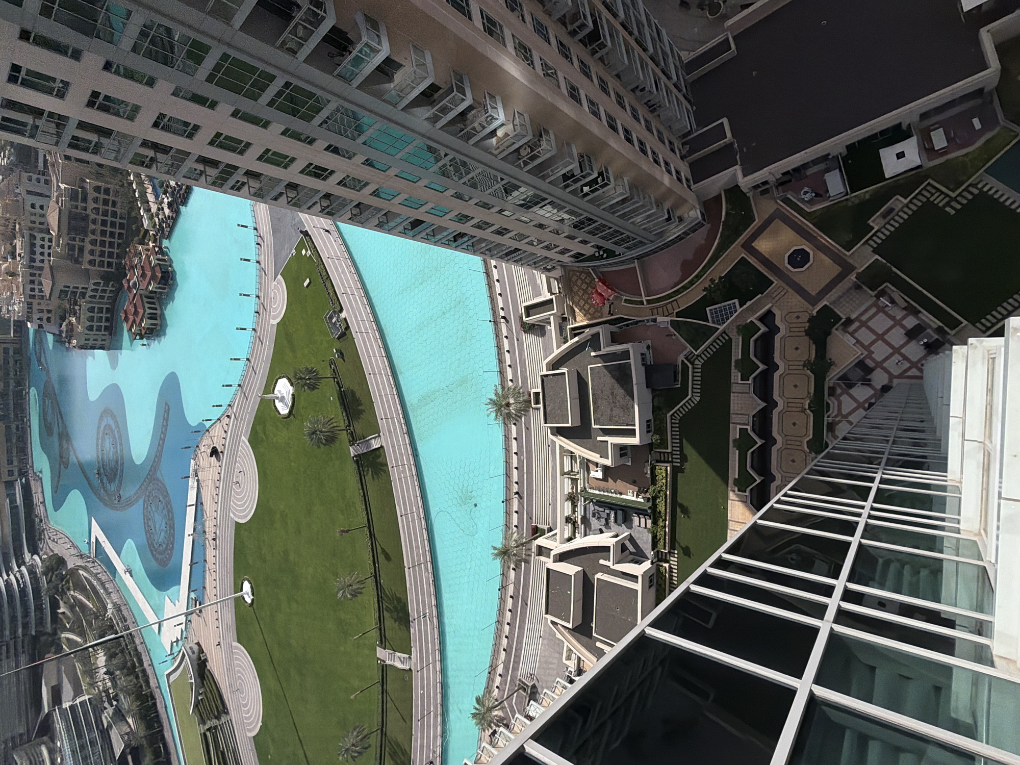 The image presents a high-angle view in Dubai, United Arab Emirates. A large, blue artificial lake featuring decorative patterns is seen on the left. Green lawns with walkways and palm trees line the waterfront. Adjacent to the green space are buildings with dark roofs and light-colored walls. Tall buildings with glass windows are on the right and upper part of the image.
