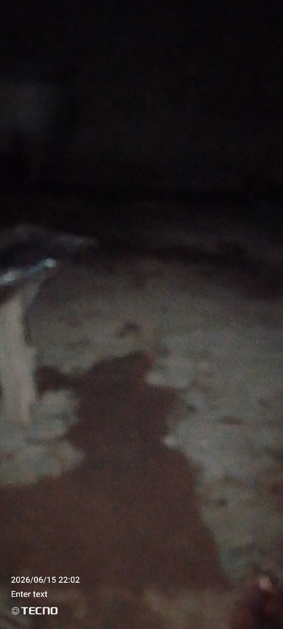 The image appears to be a photograph taken at night, possibly in Jalingo, Nigeria, given the metadata. It shows a dimly lit outdoor scene with a dark, textured ground surface. In the center of the frame, there is a prominent shadow cast on the ground, suggesting the presence of an object or person illuminated from an unseen light source. The shadow is irregular in shape, with darker and lighter areas, indicating the form is not clearly defined. There are faint, lighter patches on the ground, potentially from light reflecting off moisture or uneven surfaces. In the bottom left corner, text overlays are visible, including a date and time stamp of "2026/06/15 22:02," the phrase "Enter text," and a copyright symbol followed by "TECNO," indicating the image was captured by a TECNO device. The overall quality of the image is low due to darkness and possibly motion blur, making precise identification of specific elements challenging.