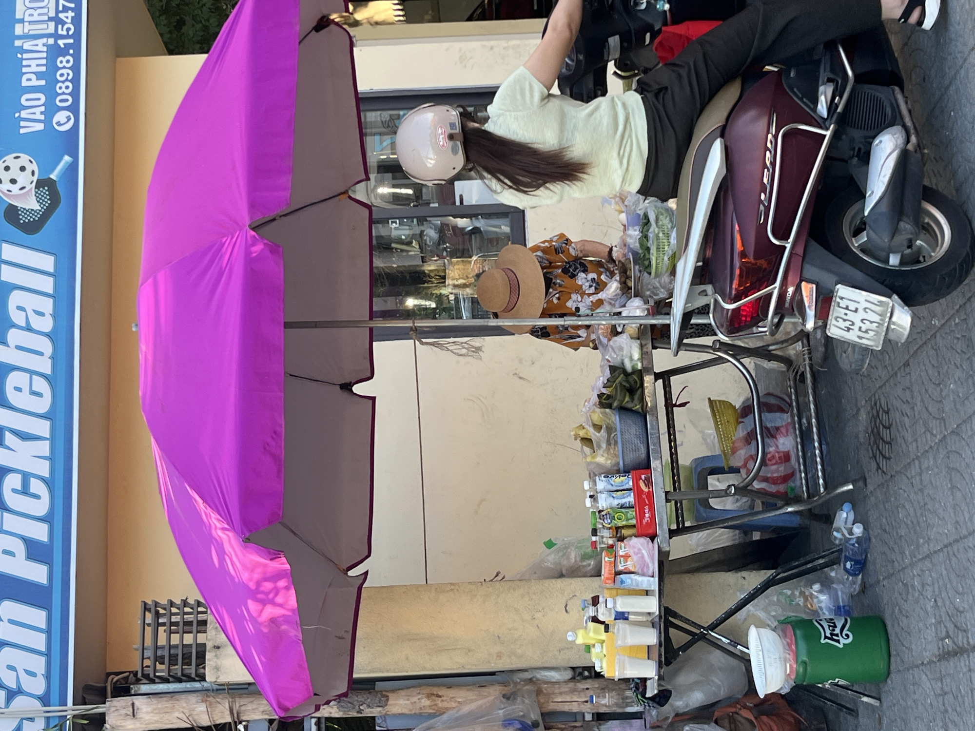 A street vendor is selling drinks and possibly snacks from a metal table under a large pink umbrella in Da Nang, Vietnam. A woman wearing a tan hat and floral shirt stands behind the table, attending to her goods. A woman wearing a light green shirt and black pants, with a white helmet, is sitting on a maroon scooter parked to the right of the vendor. The scooter's license plate reads "43-E1 153.74". The background shows a light-colored building with a blue sign at the top that reads "San Pickleball" and "VÀO PHÍA TRỌNG" with a phone number "0898.1547". The scene appears to be a sidewalk in a city, with a paved ground surface and a drain grate visible. Bottles, cups, and various items are arranged on the vendor's table.