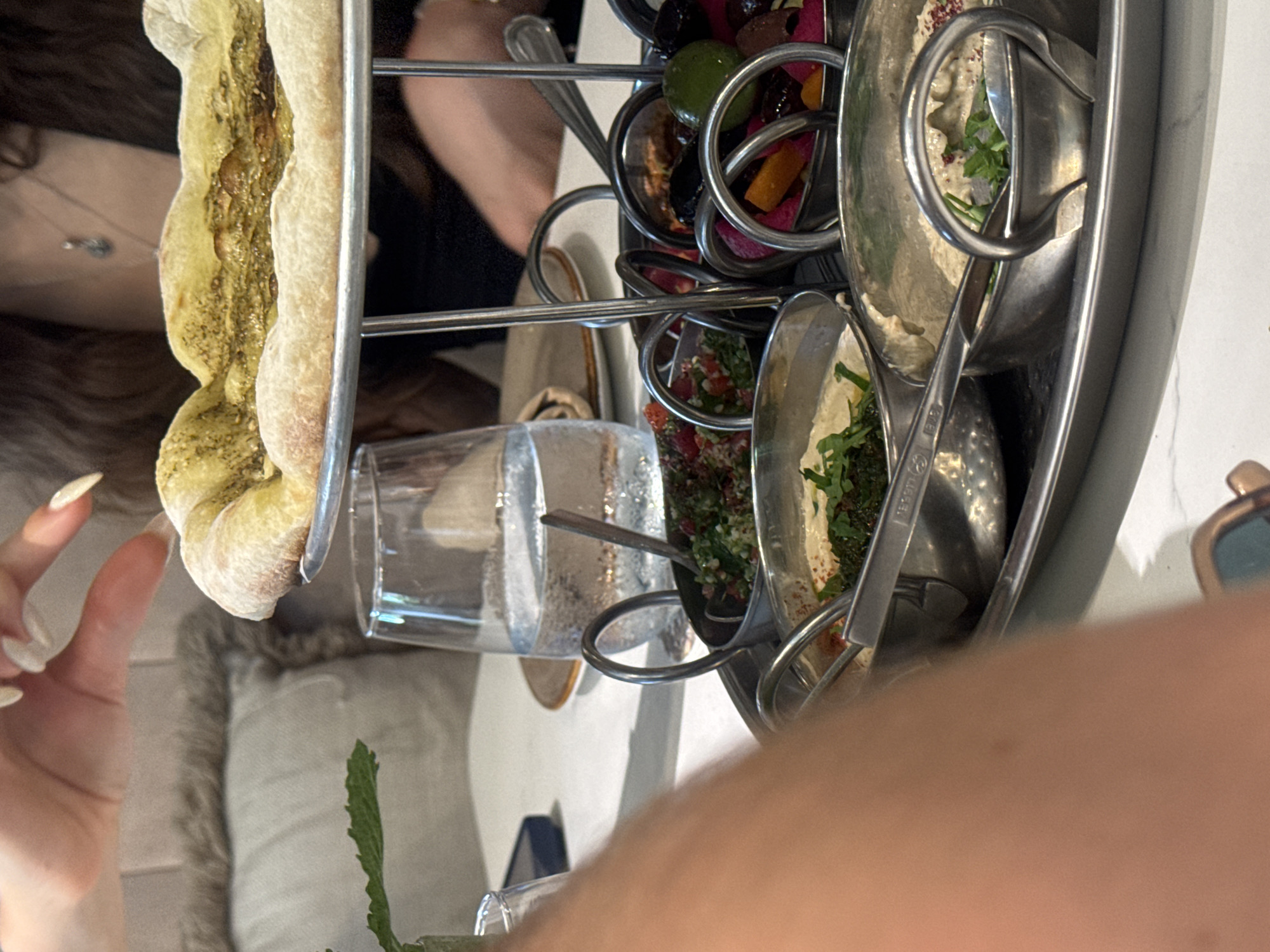 The image depicts a close-up view of a dining table in Aventura, United States of America, with a focus on a tiered serving platter of Mediterranean appetizers. A person's hands with manicured nails are visible in the upper left, reaching towards a flatbread with a green spread, possibly za'atar or pesto, which is positioned on the top tier of the platter. The flatbread is golden brown with a slightly puffed crust. Below the flatbread, the platter holds several small metal bowls filled with various dips and toppings. These include a vibrant red salad, likely a tomato and herb mix, and a creamy white dip, possibly hummus, garnished with parsley and red pepper flakes. Another bowl contains an assortment of olives, pickles, and possibly pickled turnips, with a bright green lime wedge visible. A glass of water with ice is also present on the table, next to the platter. The environment appears to be a restaurant setting, suggested by the table linens, glassware, and the background elements, including a plush cushion on a chair and the blurred figure of another diner. The overall lighting suggests an indoor daytime setting.