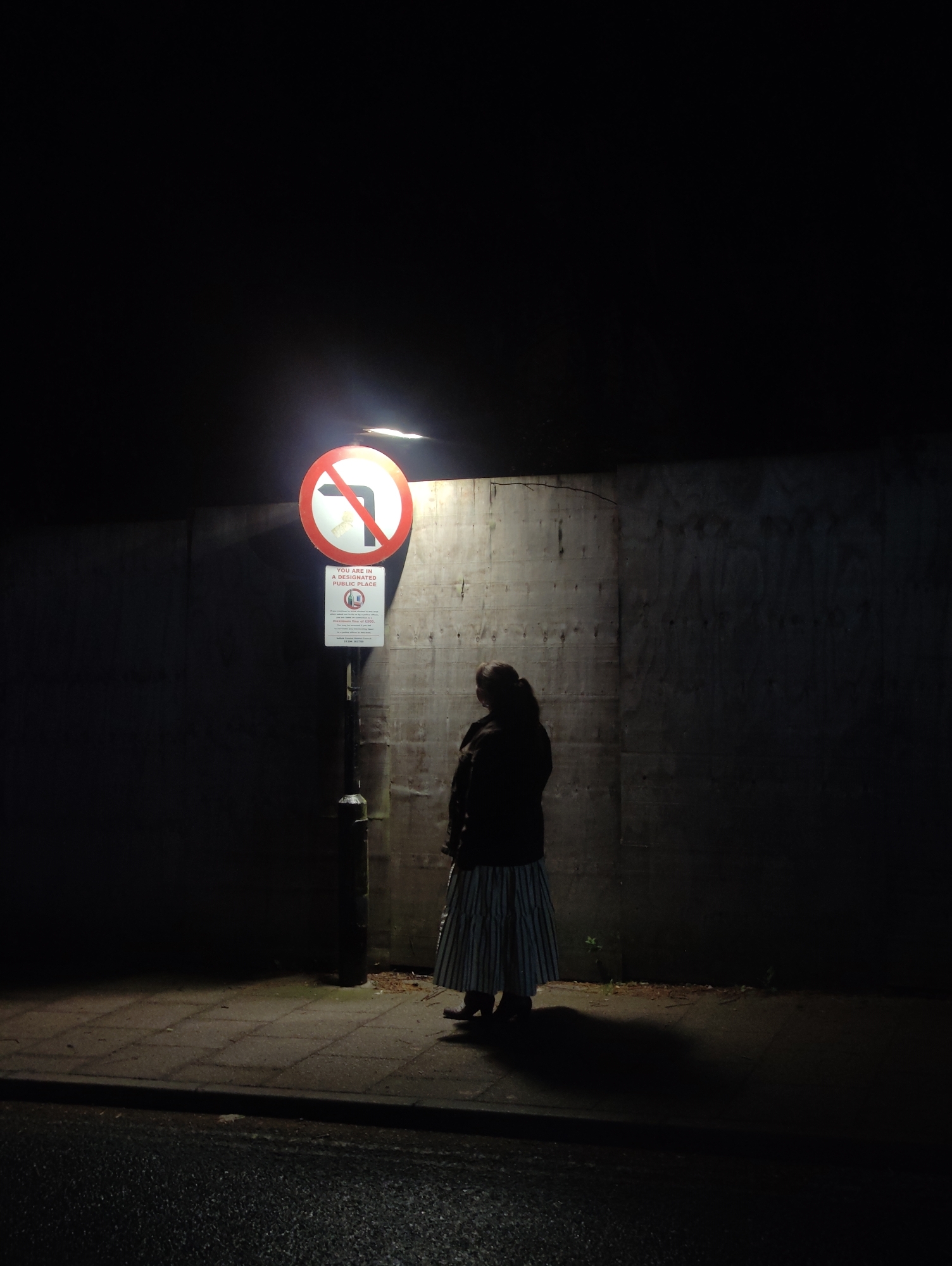 A woman stands on a sidewalk at night in East Suffolk, United Kingdom. She faces a weathered concrete wall illuminated by an overhead streetlight. A "no left turn" traffic sign is affixed to the light pole, with a smaller sign beneath indicating the location is a designated public place. The woman wears a dark jacket and a long striped skirt. Her shadow is cast on the sidewalk. The scene is darkly lit.