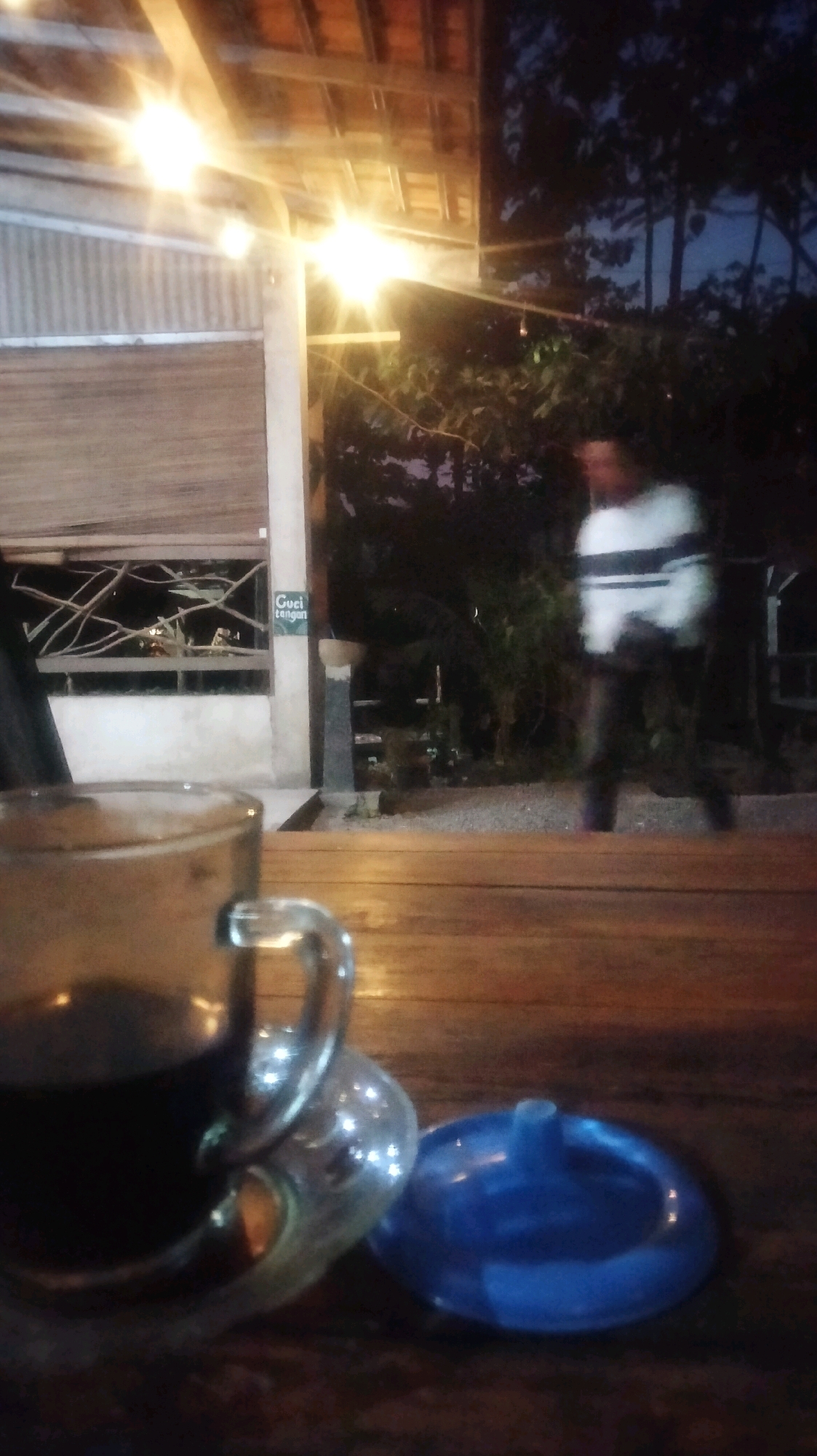 The image depicts an outdoor seating area at night, likely in Plumpang, Indonesia, with a focus on a wooden table in the foreground. On the table, a glass mug filled with a dark liquid, presumably coffee, is prominently placed to the left. Next to it, a blue ashtray rests on the table. In the background, a person wearing a white shirt with horizontal black stripes and dark pants is walking away from the camera. The setting features overhead lighting with exposed bulbs and a slatted roof structure, creating a warm, ambient glow. Beyond the seating area, there are trees and foliage, suggesting a natural or semi-natural environment. A sign reading "Cuci Tangan" (Wash Hands) is visible on a pillar to the left of the background figure. The overall atmosphere is casual and relaxed, indicative of a nighttime gathering spot.