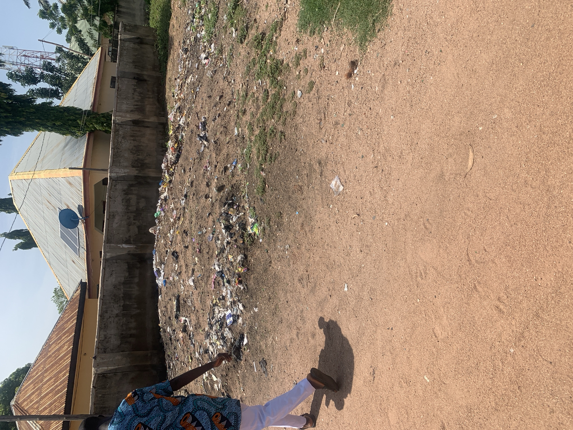 The image depicts an outdoor scene in Jauro Yinu, Nigeria, characterized by a pile of garbage situated next to a wall and some residential buildings with corrugated metal roofs. The garbage is a mix of various items, including plastic bags, bottles, and other debris, with some patches of dry grass interspersed within it.
In the foreground, a person is visible from the knees down, walking away from the camera on a dirt path. They are wearing white pants and dark sandals, and a colorful, patterned shirt is partially visible. The person's shadow is cast on the path to their right. The ground is sandy and uneven, with scattered small stones and debris.
Behind the garbage pile, several buildings are partially visible. One building has a light yellow facade and a light gray corrugated metal roof, with a blue satellite dish and solar panels on top. Another building to the left has a similar corrugated metal roof, but appears to be older and more weathered. In the background, trees and communication towers can be seen against a clear blue sky, suggesting a sunny day.
The overall scene conveys a sense of neglect and environmental concern due to the presence of the large amount of waste. The time of day appears to be daytime, given the clear sky and the shadows cast. No text is visible in the image.
