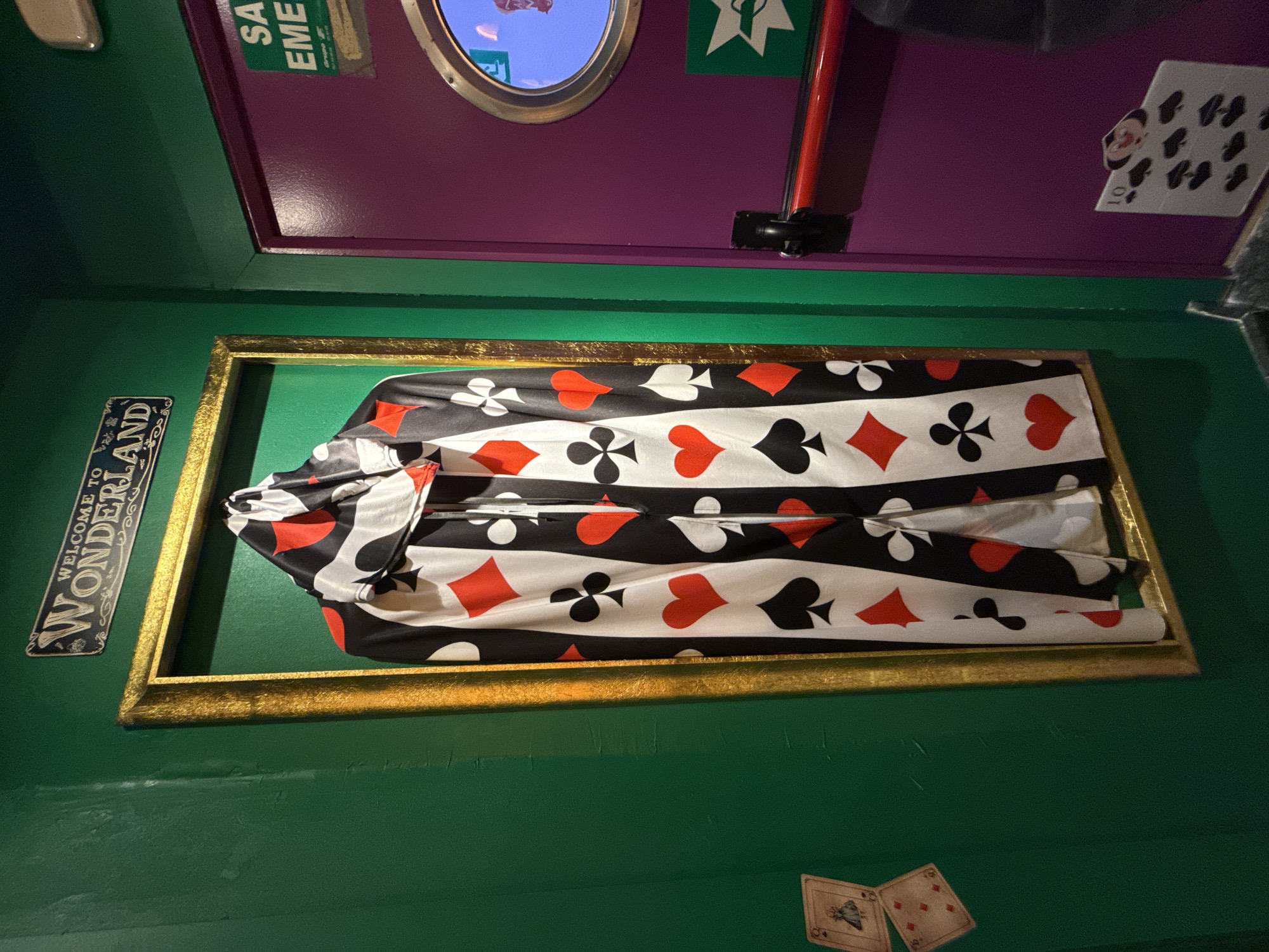 This image depicts a decorative display likely related to the theme of Alice in Wonderland, possibly found in an entertainment venue or themed restaurant. The room has green walls, and above the display is a purple-colored wall with a porthole-style window. The display features a golden rectangular frame containing a bundle of fabric with a pattern of playing card suits (hearts, spades, diamonds, clubs). To the side of the frame hangs a sign that says "Welcome to Wonderland." Additionally, playing cards are stuck to the wall near the frame and the porthole. While not explicitly confirmed, this scene is potentially located in Madrid, Spain, based on the image context. The lack of people and actions suggests a static display meant for visual entertainment.
