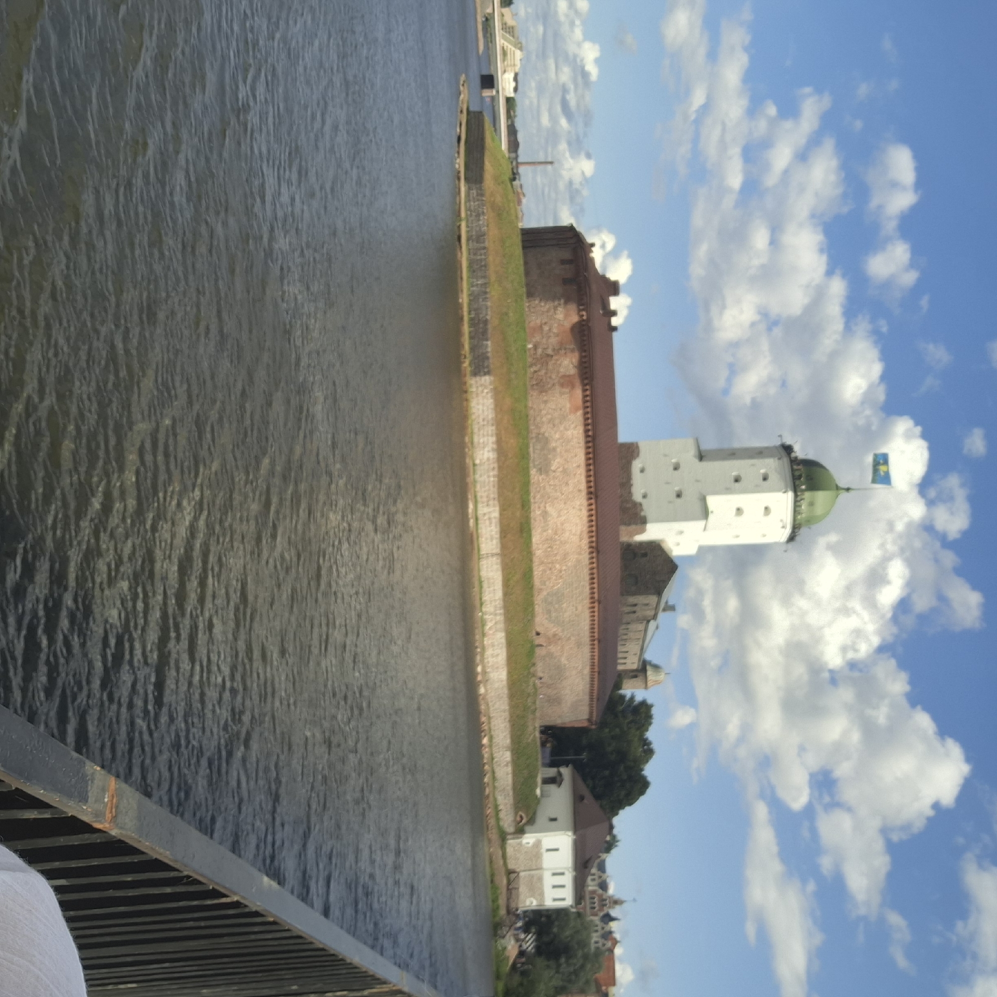 The image shows Vyborg Castle, a medieval castle located on the Karelian Isthmus in Vyborg, Russia. The castle is situated on a small island in Vyborg Bay and is a prominent landmark in the city. The image captures the castle from across the bay, with the water in the foreground and the castle complex dominating the background.
The castle consists of a main tower, known as St. Olaf's Tower, which is tall and white with a green dome and a flag flying at its peak. Surrounding the tower is a large, red-tiled wall with several smaller buildings integrated into the complex. The architecture appears to be a mix of medieval and later additions.
The environment depicted is a bright, sunny day with a blue sky dotted with fluffy white clouds. The water is dark and appears slightly rippled. In the distance, other buildings and greenery can be seen on the mainland, suggesting an urban or semi-urban setting. A black metal railing is visible in the lower right corner of the image, with part of a person's shoulder and white clothing in the very bottom right. The overall impression is of a historic and picturesque location.