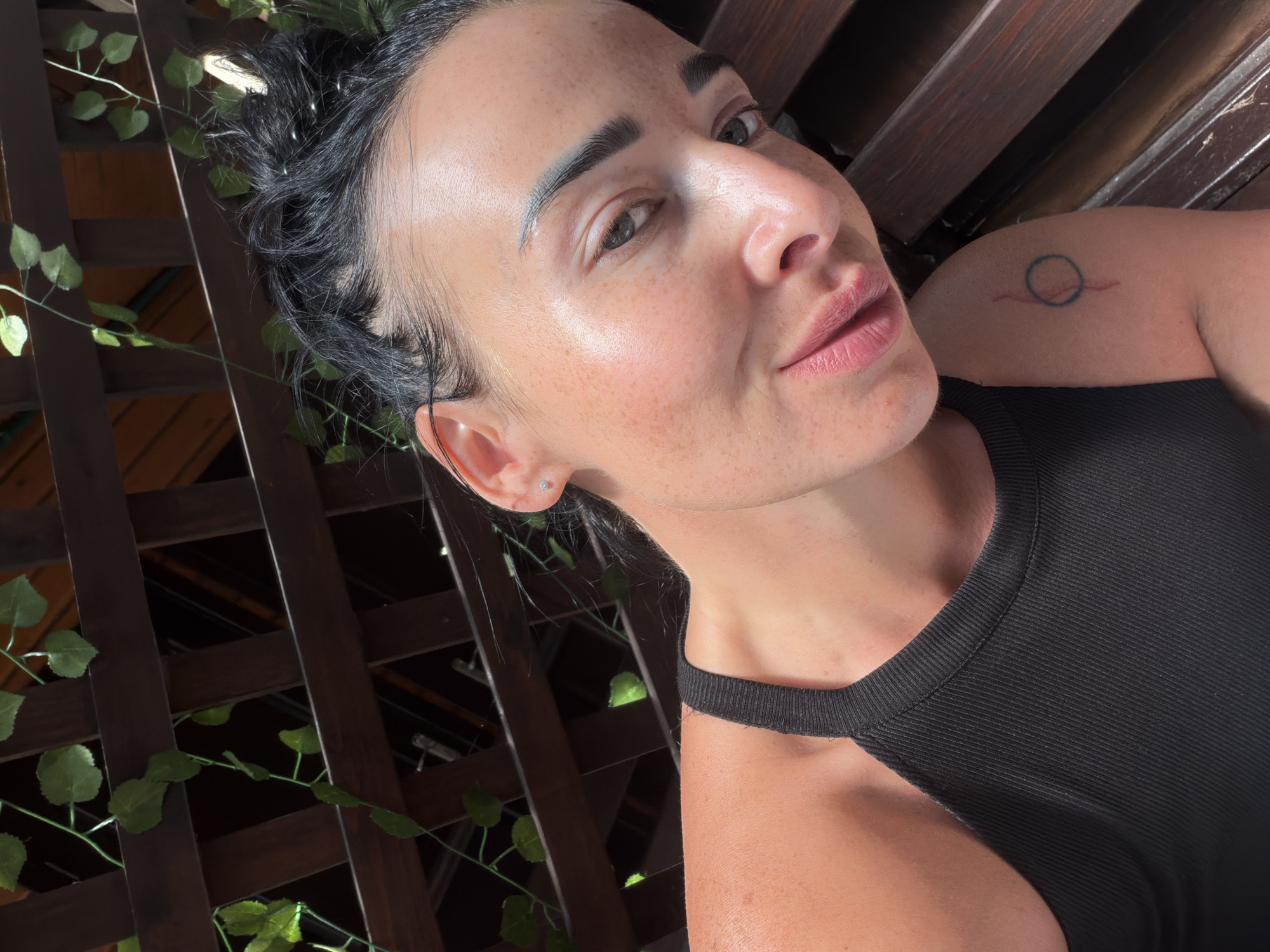 The media file is a close-up, angled selfie of a woman with dark hair pulled back. She is outdoors, with a wooden lattice structure and greenery visible in the background, suggesting a garden or patio setting. The lighting indicates it is daytime, possibly late afternoon or early evening, with soft sunlight casting shadows.
The woman has a light dusting of freckles across her nose and cheeks. She is wearing a black ribbed tank top with a cutout detail at the shoulder. A small, circular tattoo with a line through it is visible on her left shoulder. She has a small stud earring in her left ear. Her expression appears relaxed and engaging. The overall mood is casual and natural. The location context provided is Sochi, Russia.