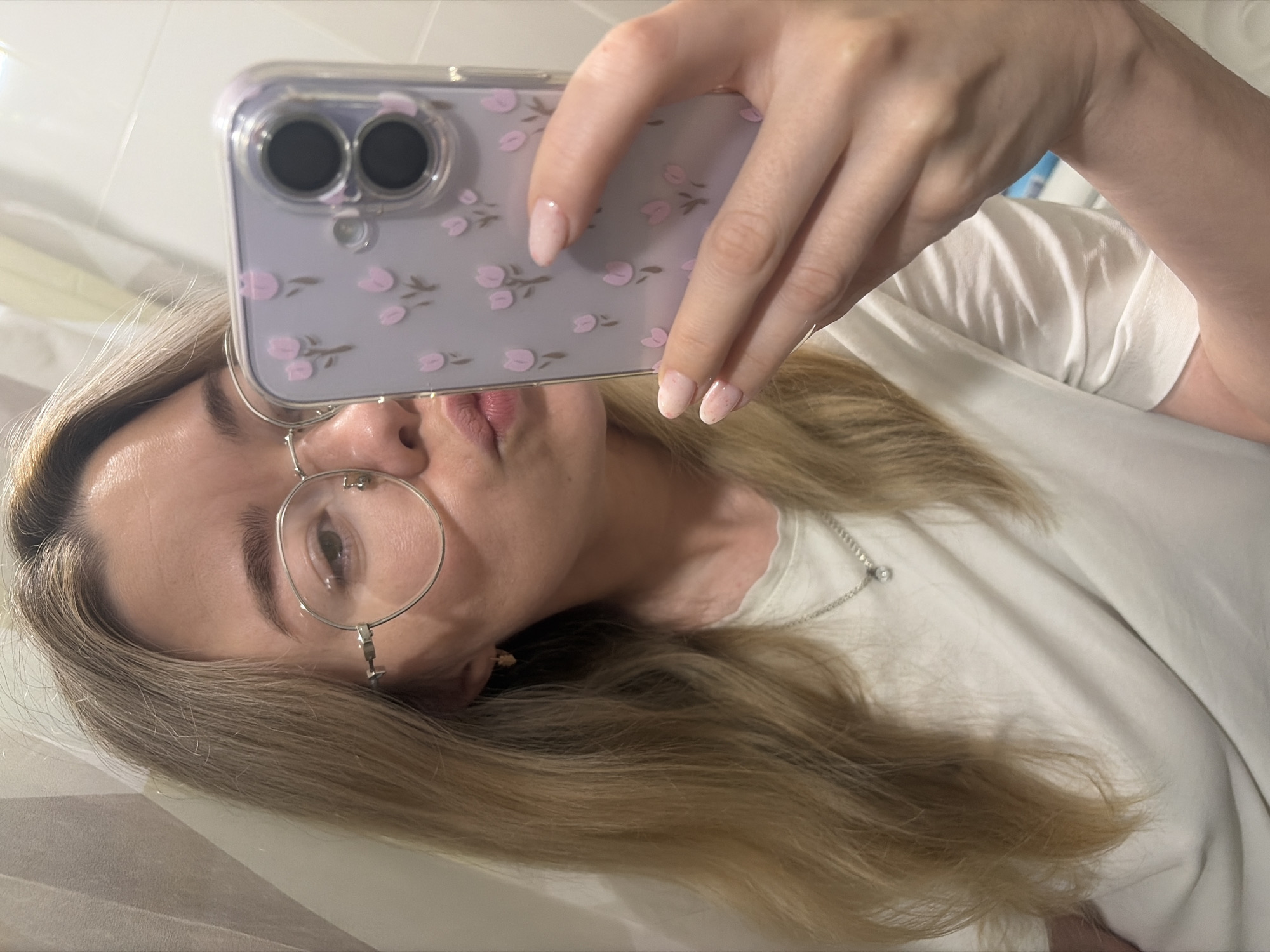 A woman is taking a selfie with a smartphone in a bathroom. The phone has a light purple case decorated with pink flower motifs. She has light blonde hair, wears round glasses, and a white t-shirt. A silver necklace is visible around her neck. The background shows white tiled walls and a white countertop or sink. The location is Chaikovsky, Russia.