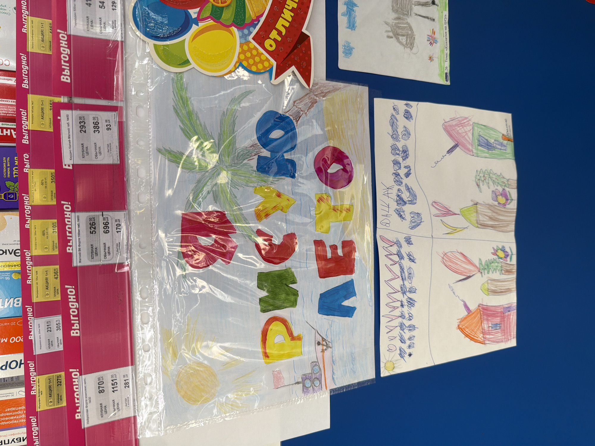 This image captures an indoor scene, likely in a commercial establishment within Leningradskaya, Russia, given the Cyrillic text and location context. The primary subjects are several children's drawings displayed on a blue surface, and numerous vertically arranged product labels on a retail rack.
The children's artwork includes a prominent drawing featuring a palm tree, sun, and water, with colorful block letters spelling "РАССВЕТ" (Sunrise) across it. Other drawings depict childlike figures, landscapes, and animals. One drawing has text "Тема: «Козлятки на дугу»" (Theme: "Goats/Kids on the Arc/Bow"), indicating the subject matter. Another drawing is signed "ДАШЕ AK" (Dasha AK), likely the artist's name. Some drawings are covered by a transparent plastic sheet.
Parallel to these displays, a retail rack holds various product labels, predominantly red and white, advertising what appear to be pharmaceutical or health-related items. The labels prominently feature the word "Выгодно!" (Profitable! or Good Deal!) and display product names such as "Ибупрофен," "Нейроспан Форте," "Магний В6 Форте," and "Кардио Калий-Магний." Details like "КЛУБНАЯ ЦЕНА" (Club Price), "ОБЫЧНАЯ ЦЕНА" (Regular Price), and promotions like "АКЦИЯ! 1+1" (Action! Buy one get one free) are visible, along with prices in rubles. A decorative cutout with the partially visible word "отлич" (likely part of "Отлично!" meaning Excellent!) is affixed near the top.
The overall scene suggests a retail environment that incorporates a community or children's art display, possibly in a pharmacy or supermarket health section, reflecting local engagement alongside commercial activity. The image is rotated, showing the labels and drawings oriented vertically.