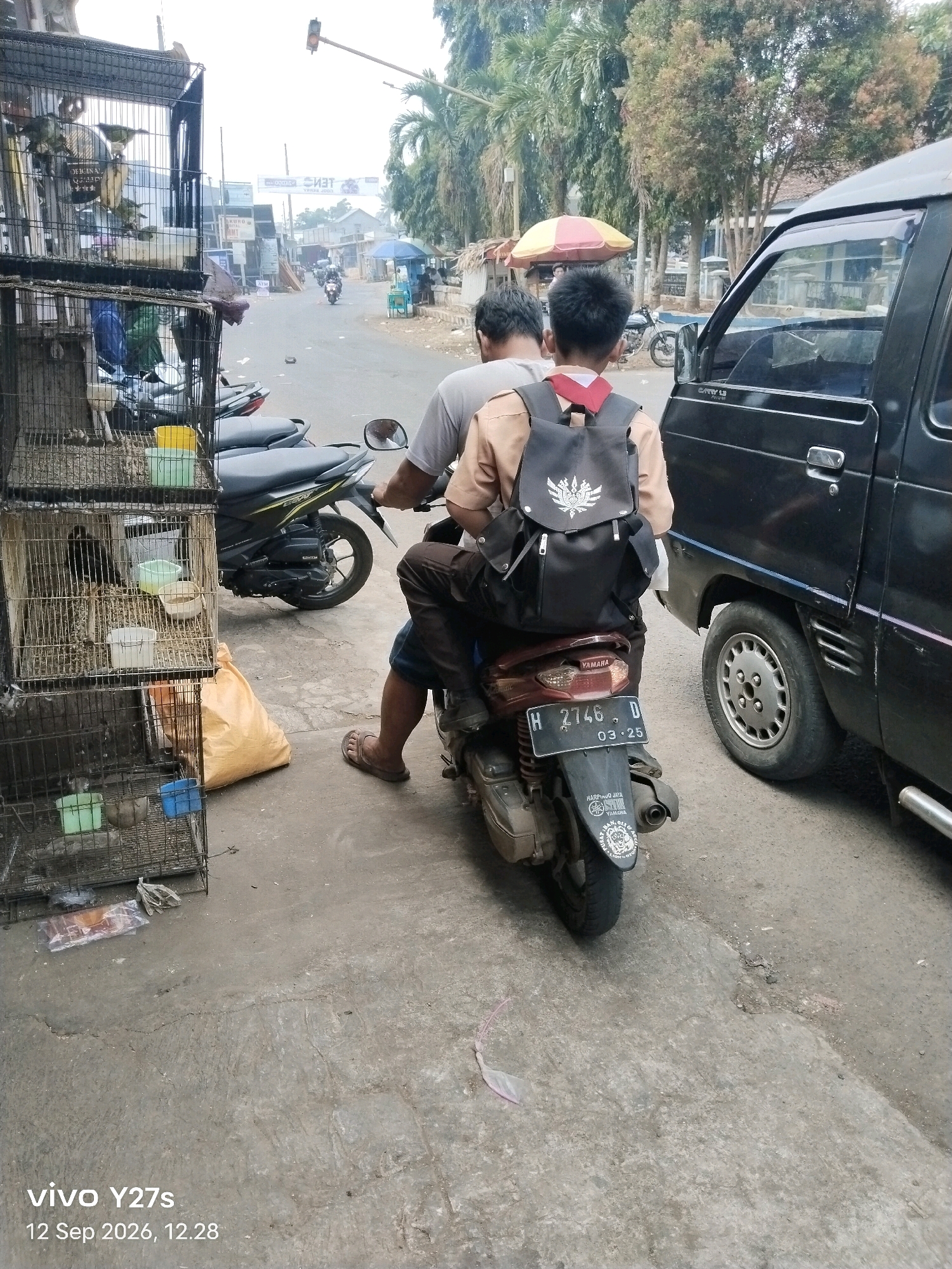In Trimulyo, Indonesia, on September 12, 2026, at 12:28, two individuals are seated on a Yamaha motorcycle. The person in the rear, a young male wearing a brown uniform and a black backpack, is facing away from the camera. The individual in front, whose face is not visible, is wearing a grey shirt. The motorcycle has a license plate reading "H 2746 D" and displays a "03-25" sticker. To the left of the motorcycle, multiple bird cages are stacked, containing birds. A large yellow bag is placed on the ground next to the cages. A black van is parked to the right of the motorcycle. The scene appears to be along a street with a paved surface, lined with trees and buildings in the background. Traffic signals and various signs are visible in the distance.