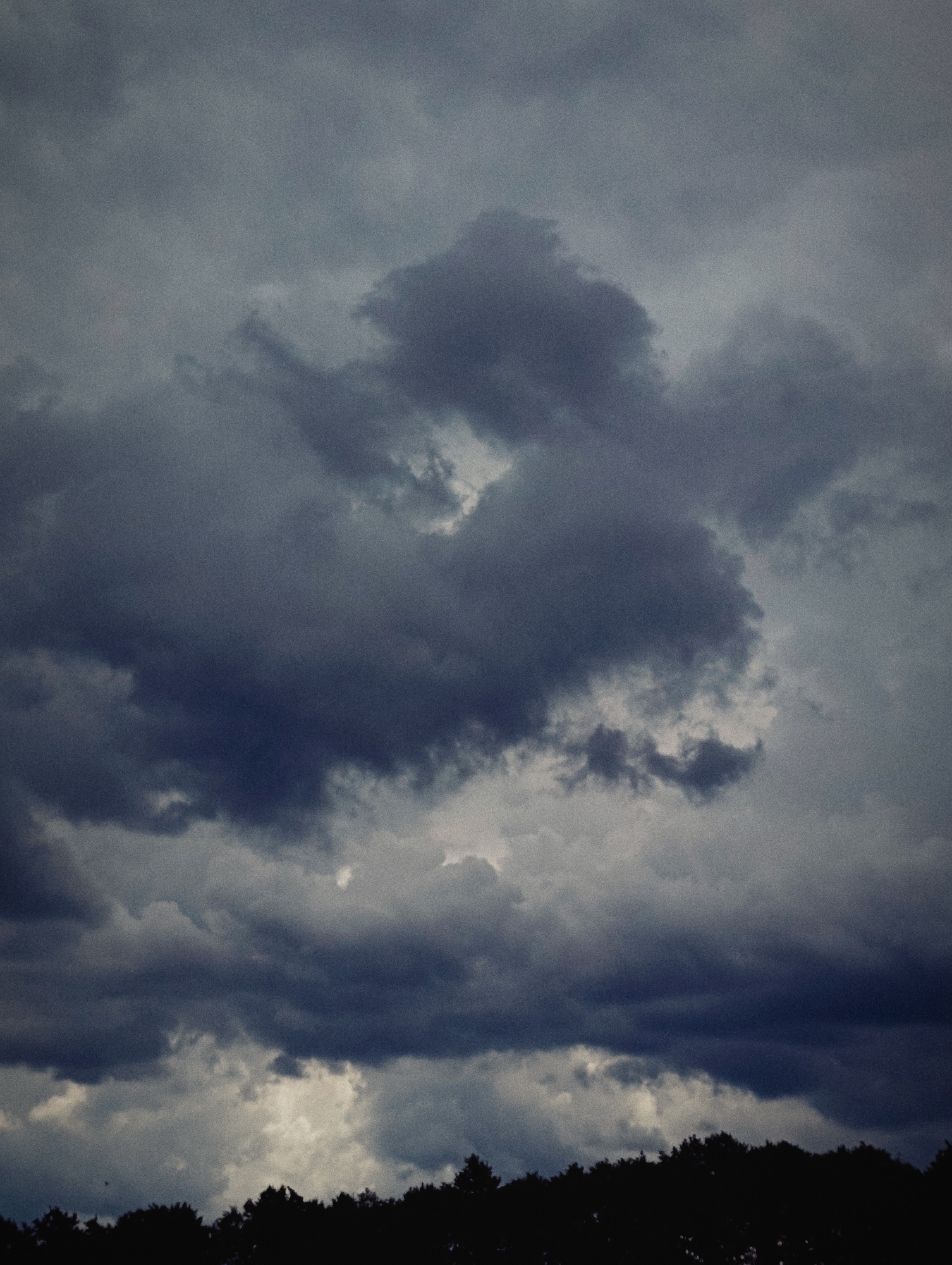 The image depicts a sky filled with dark, ominous clouds, suggesting an impending storm or overcast weather. A silhouette of a dense line of trees forms the bottom edge of the frame, creating a stark contrast against the turbulent sky. The scene is devoid of any human presence or visible urban structures, making it impossible to ascertain a specific location within Minsk, Belarus, or any city for that matter. The overall impression is one of natural, dramatic atmospheric conditions.
