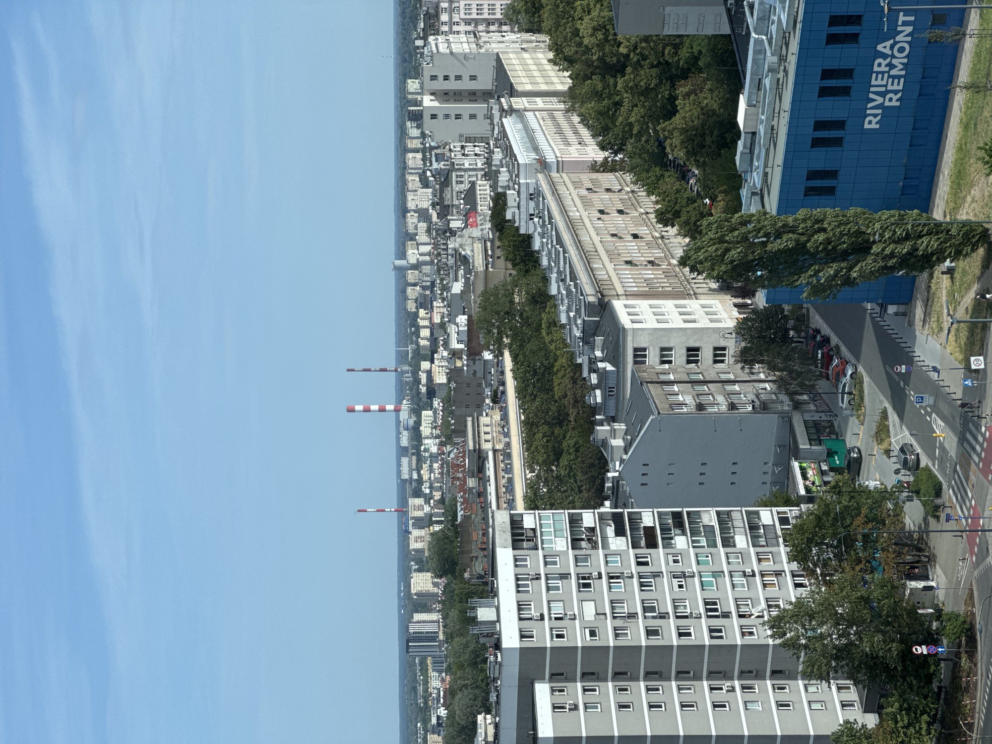 This is a high-angle, daytime, vertical shot of a cityscape in Warsaw, Poland. The scene is dominated by a sprawling urban environment with numerous buildings of varying heights and architectural styles, interspersed with green trees.
In the foreground, a tall, modern building with a distinctive blue facade stands out. White text on the blue building reads "RIVIERA REMONT". Adjacent to this is a gray, multi-story residential building with many windows. Further into the city, a long stretch of apartment buildings with a repeating pattern of windows can be seen. In the distance, two tall smokestacks with red and white stripes punctuate the skyline against a clear blue sky.
The image captures a lively urban setting. Cars are visible on the roads, and pedestrian crossings indicate active city life. The bright daylight suggests it is likely midday. The overall impression is one of a densely populated urban area on a clear day. There are no people clearly visible in the frame, but the presence of cars and buildings implies human activity.